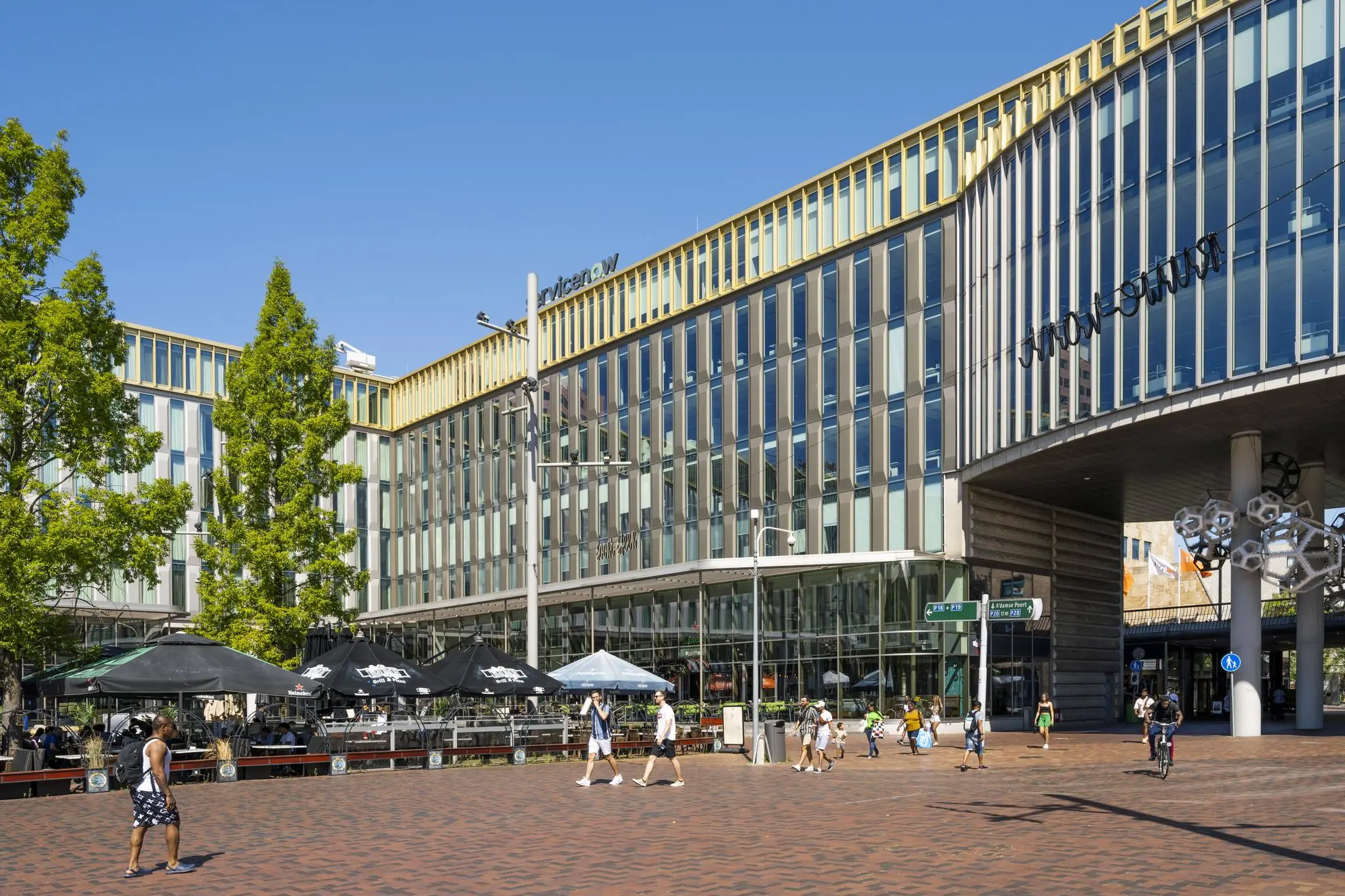 Moderne kantoorgebouwen en horecaterrassen op het Hoekenrodeplein in Amsterdam-Zuidoost met passerende voetgangers en fietsers.