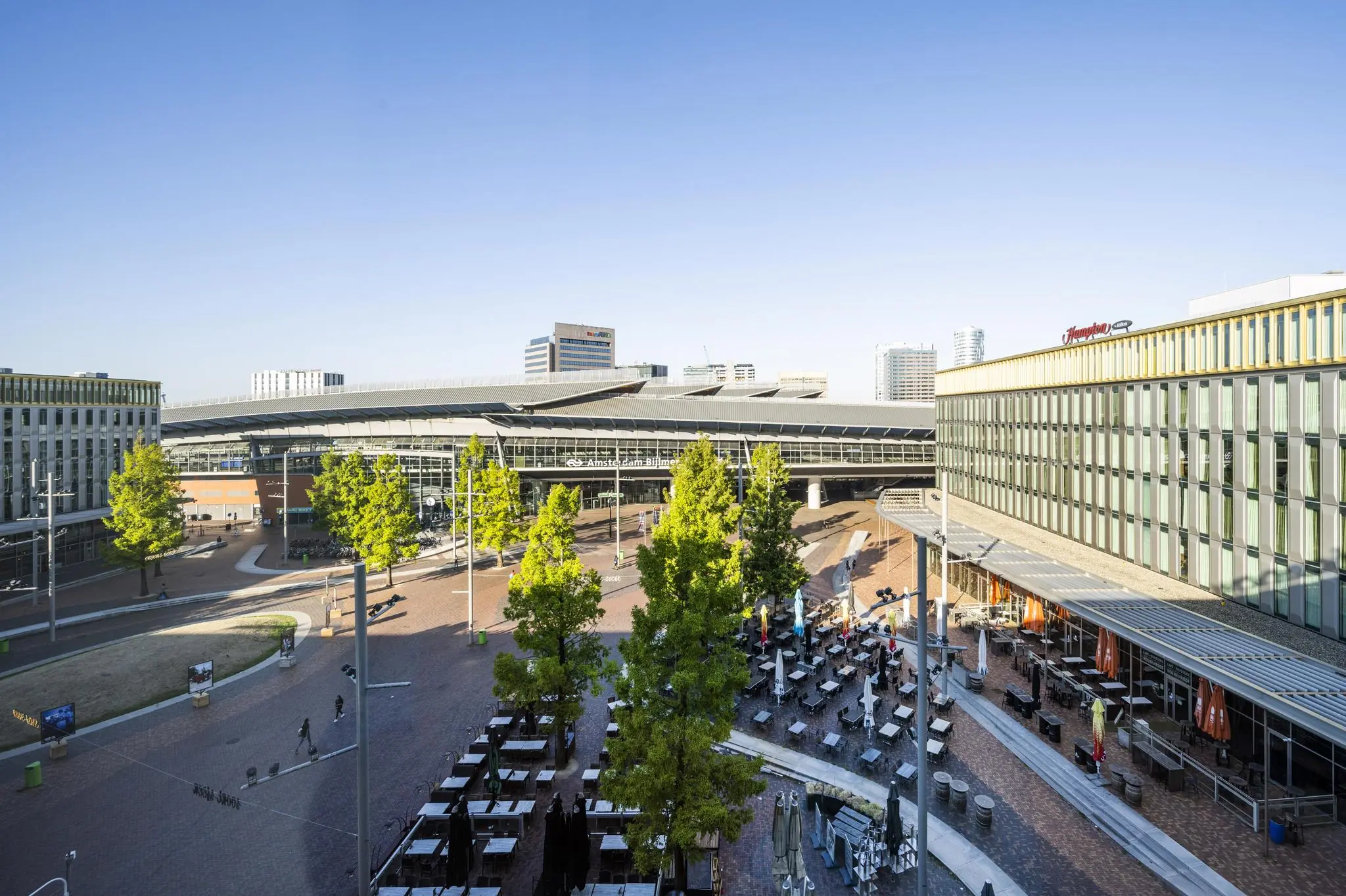 Het stationsplein bij station Amsterdam Bijlmer ArenA met horecaterras, omliggende gebouwen en bomen onder een heldere blauwe lucht.