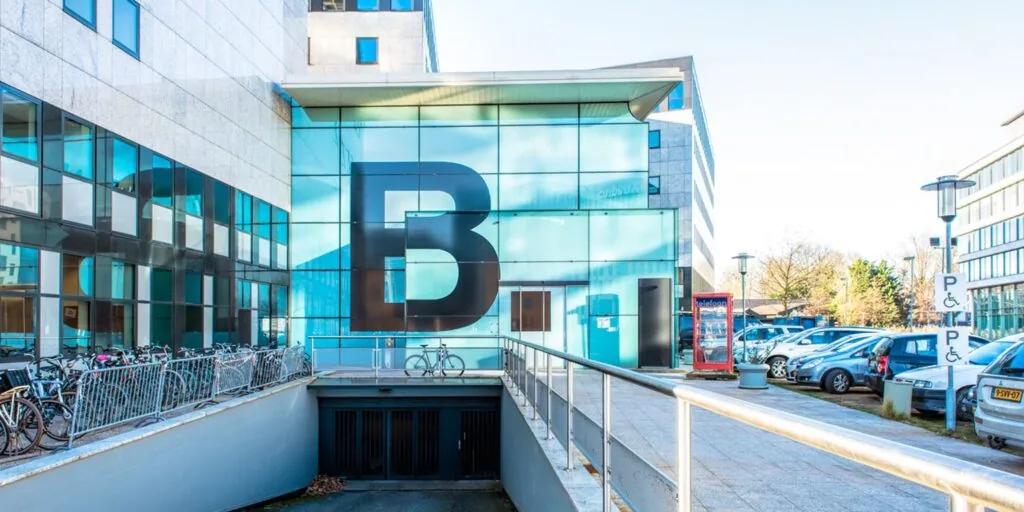 Glazen gevel van gebouw B aan het John M. Keynesplein met parkeerplaats en fietsenrek op de voorgrond.