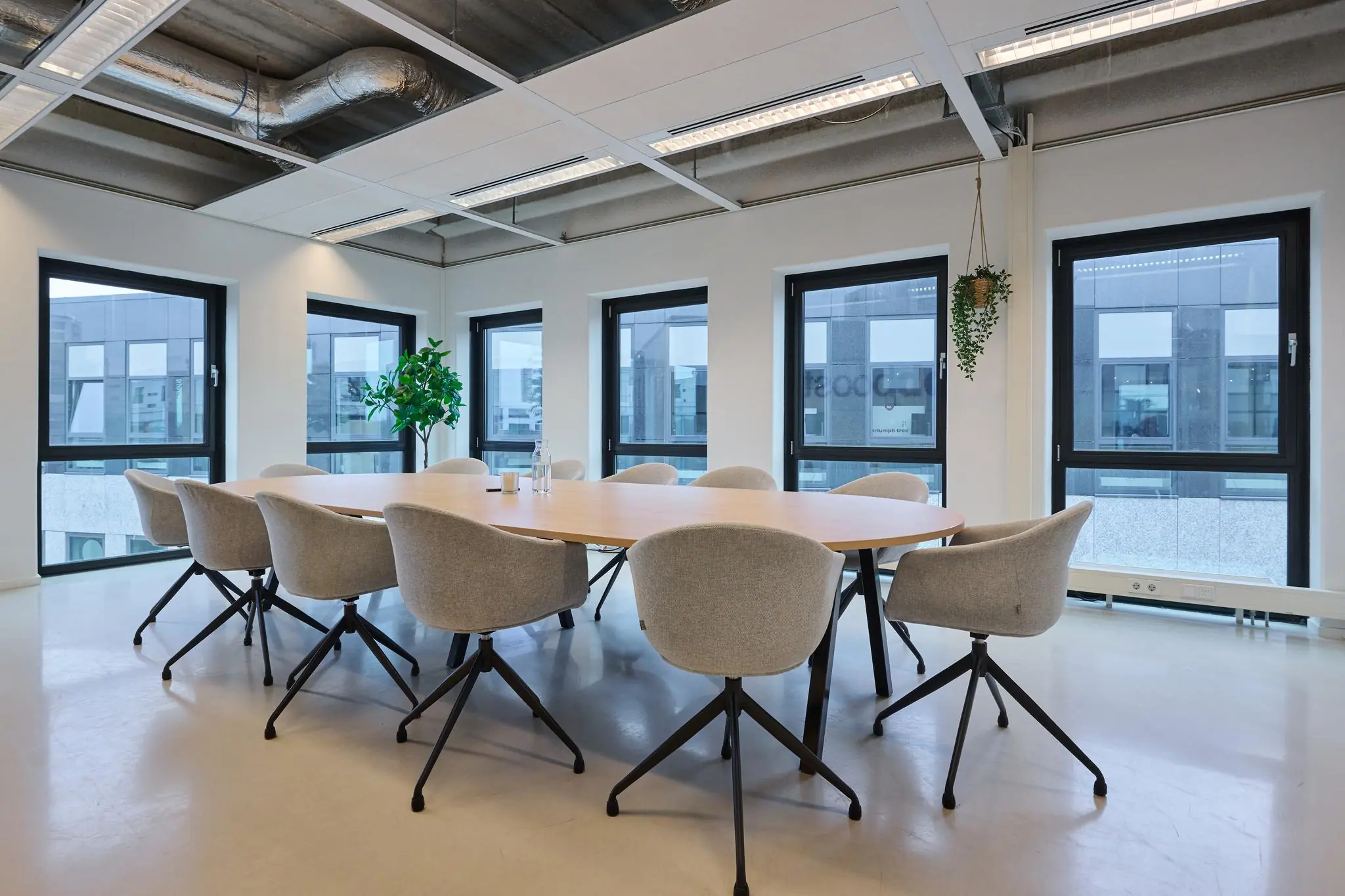 Vergaderruimte aan het John M. Keynesplein met ovale houten tafel, grijze stoelen en grote ramen met uitzicht op moderne kantoorgebouwen.