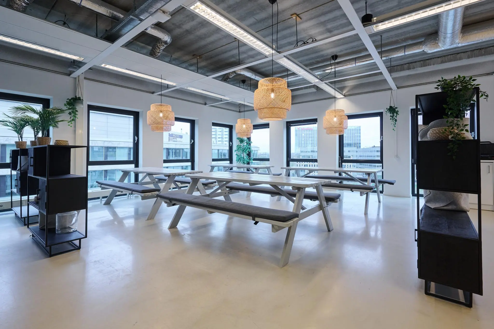 Moderne kantineruimte met houten picknicktafels, hanglampen en zicht op kantoorgebouwen aan het John M. Keynesplein.