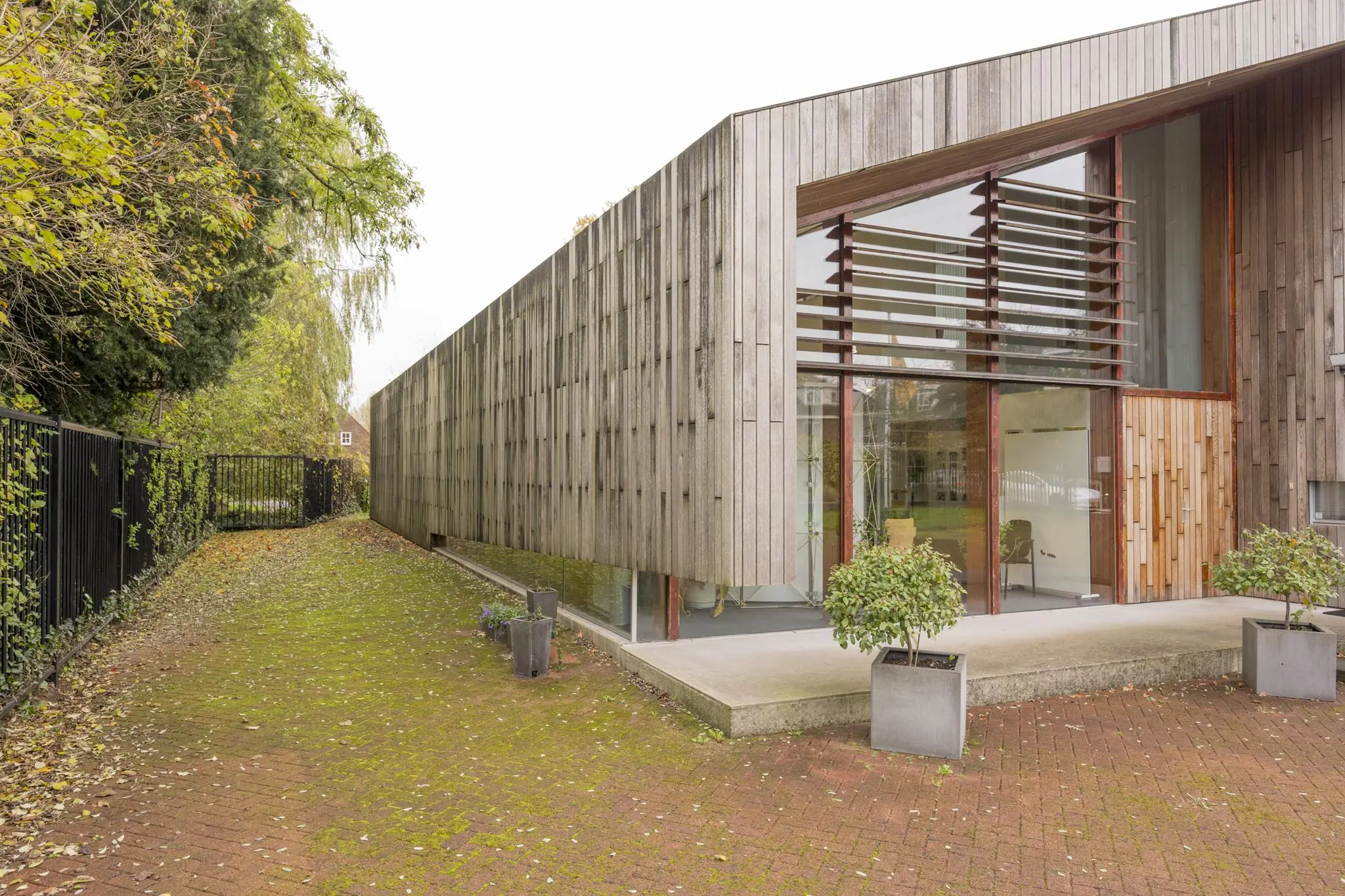 Modern houten gebouw aan de Vogelvlinderweg met grote ramen en plantenbakken op een met mos begroeide bestrating.
