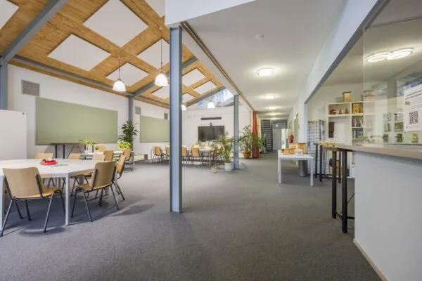 Ruime en moderne gemeenschappelijke ruimte in het gebouw aan de Vogelvlinderweg, ingericht met tafels, stoelen, planten en een presentatiehoek.