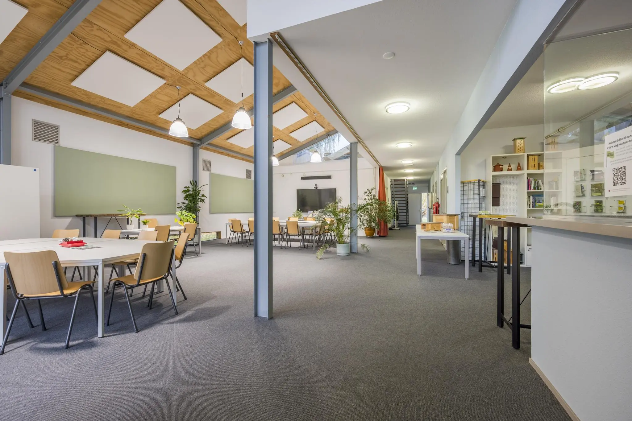 Ruime en moderne gemeenschappelijke ruimte in het gebouw aan de Vogelvlinderweg, ingericht met tafels, stoelen, planten en een presentatiehoek.