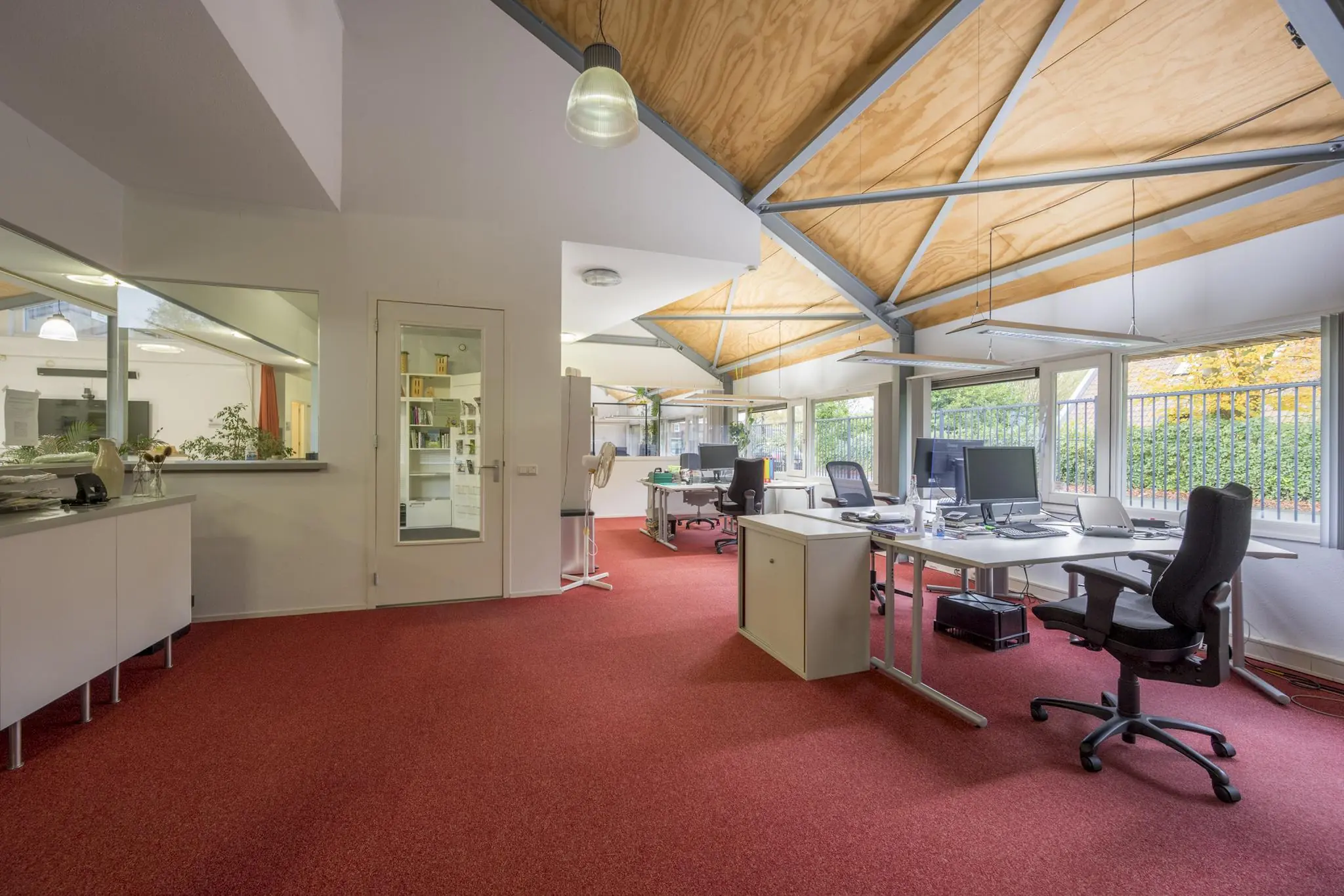 Modern kantoorinterieur aan de Vogelvlinderweg met rode vloerbedekking, schuine houten plafonds en werkplekken bij grote ramen.