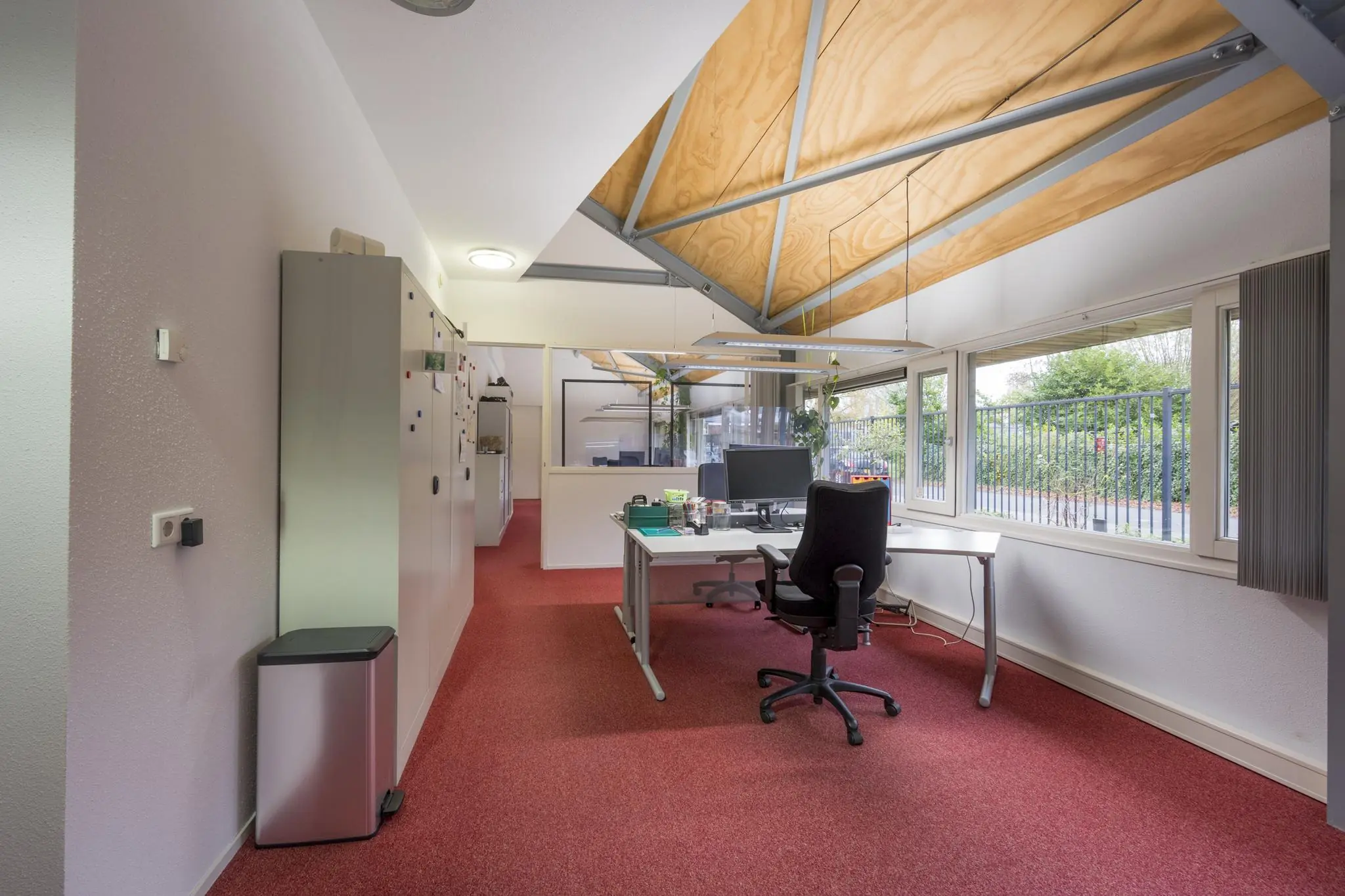 Moderne kantoorruimte aan de Vogelvlinderweg met een rood tapijt, schuine houten plafondpanelen en een bureau met computer bij het raam.