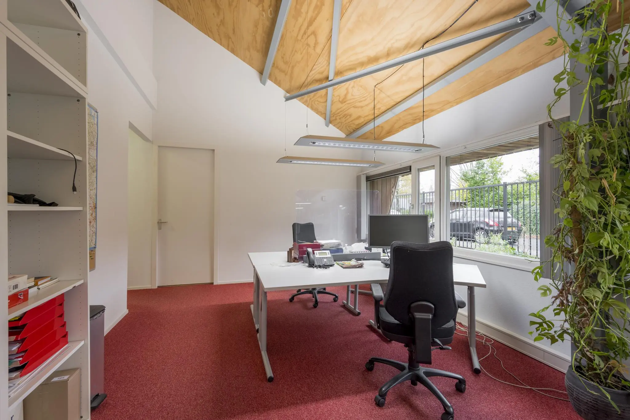 Modern ingericht kantoor met rood tapijt, houten plafond en uitzicht op een parkeerplaats via grote ramen.