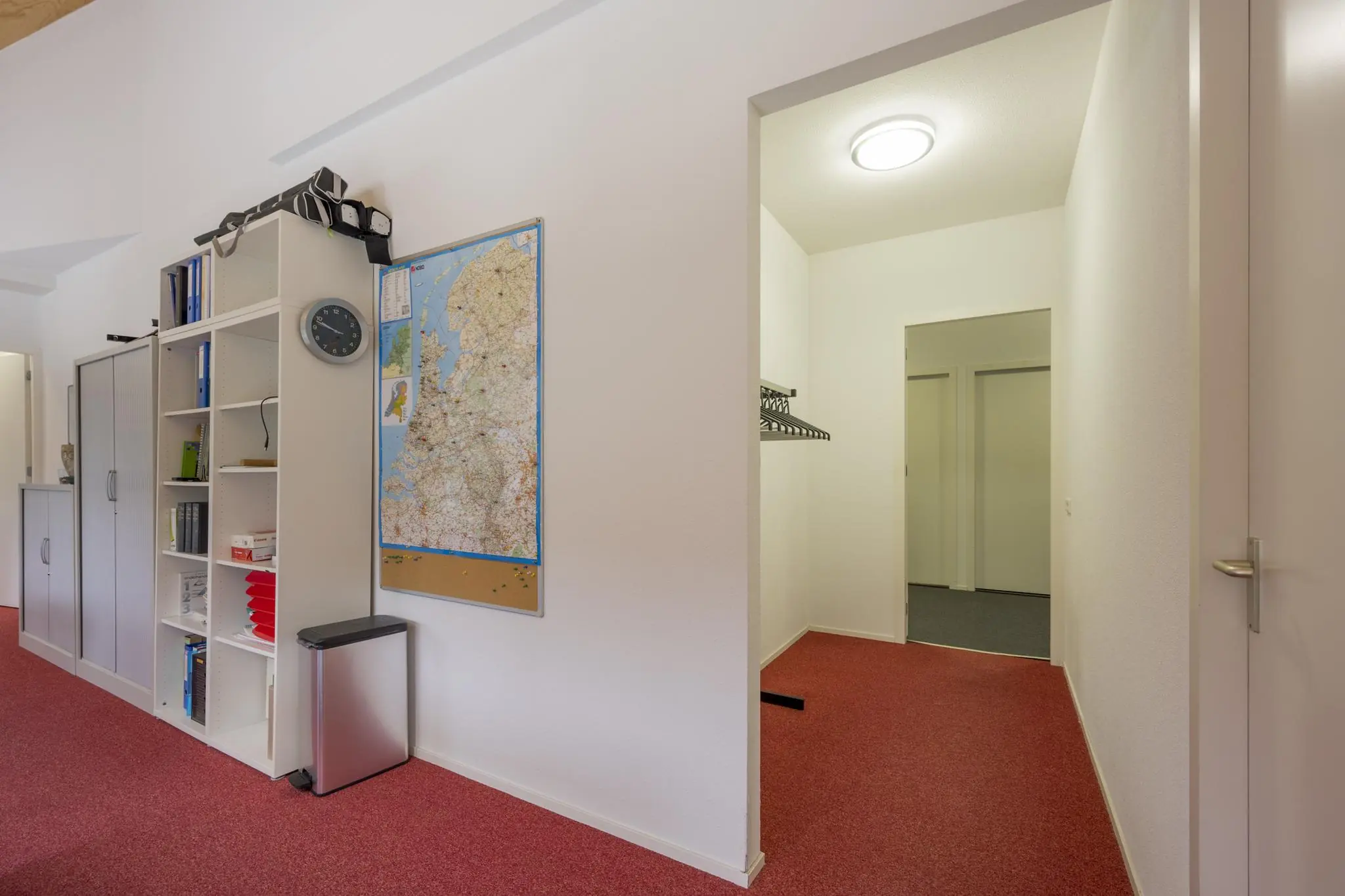 Kantoorruimte met rode vloerbedekking, een open kast, een landkaart van Nederland en een gang met kapstok.