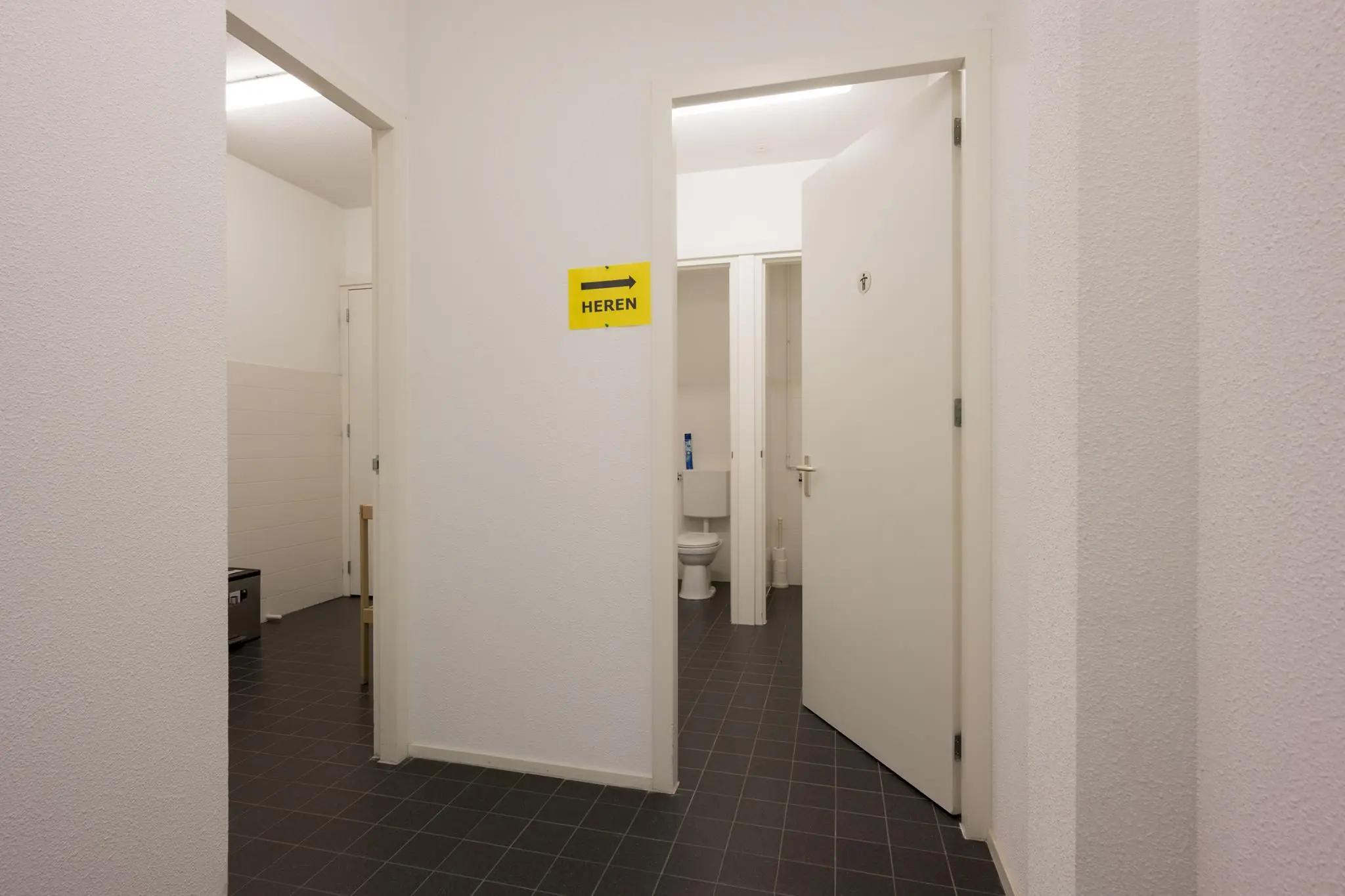 Ingang van een heren-wc met open deur en zicht op een toiletcabine in een strak, wit betegeld interieur.