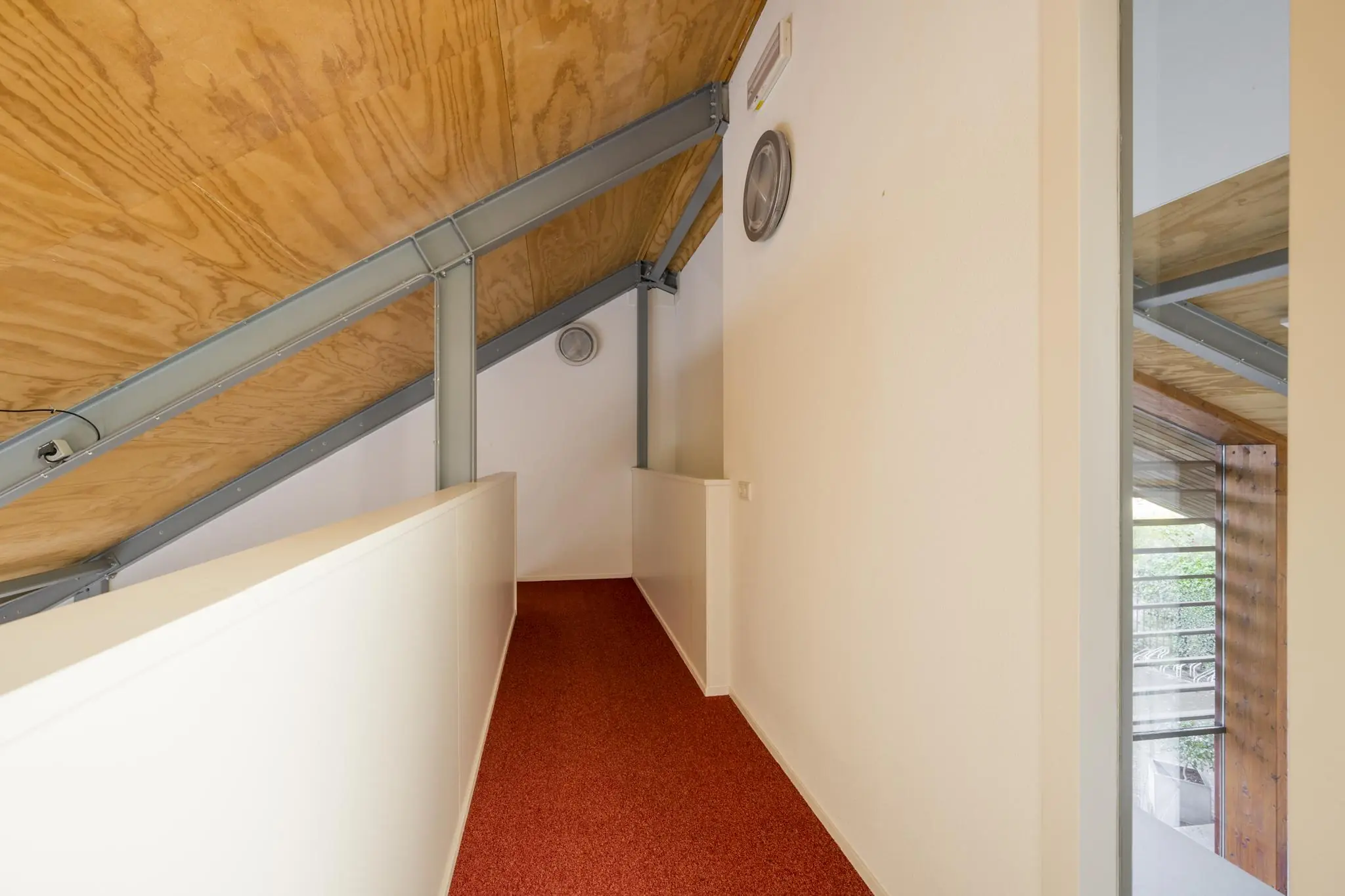 Smalle gang met rood tapijt, schuine houten plafondpanelen en metalen draagbalken in een modern huis aan de Vogelvlinderweg.