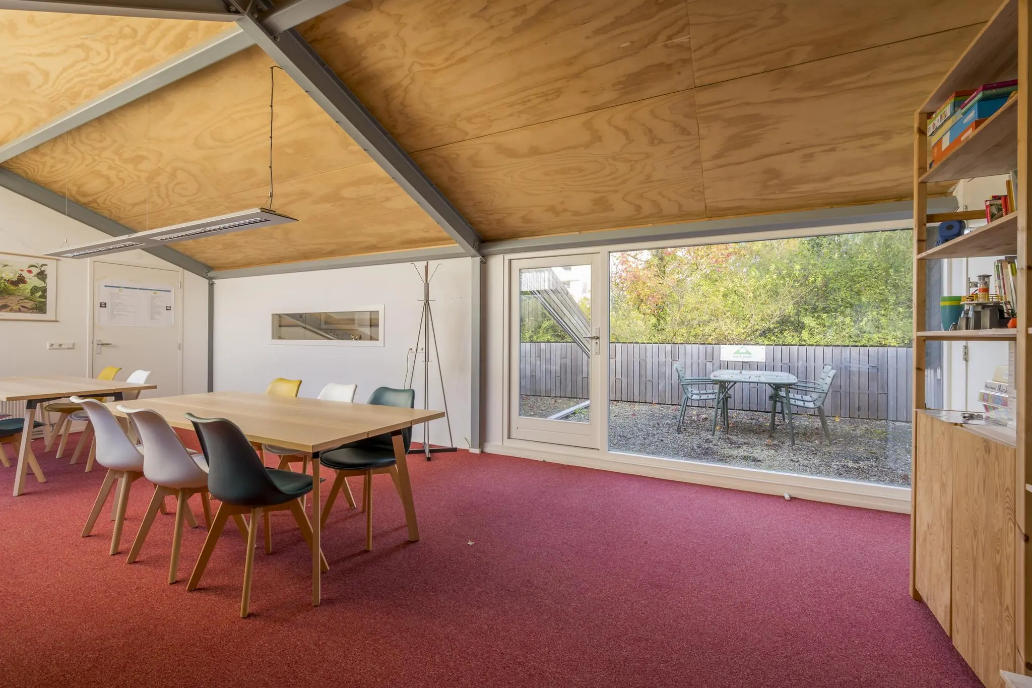 Vergaderruimte aan de Vogelvlinderweg met houten plafond, rood tapijt, kleurrijke stoelen en uitzicht op een buitenterras met zitgelegenheid.