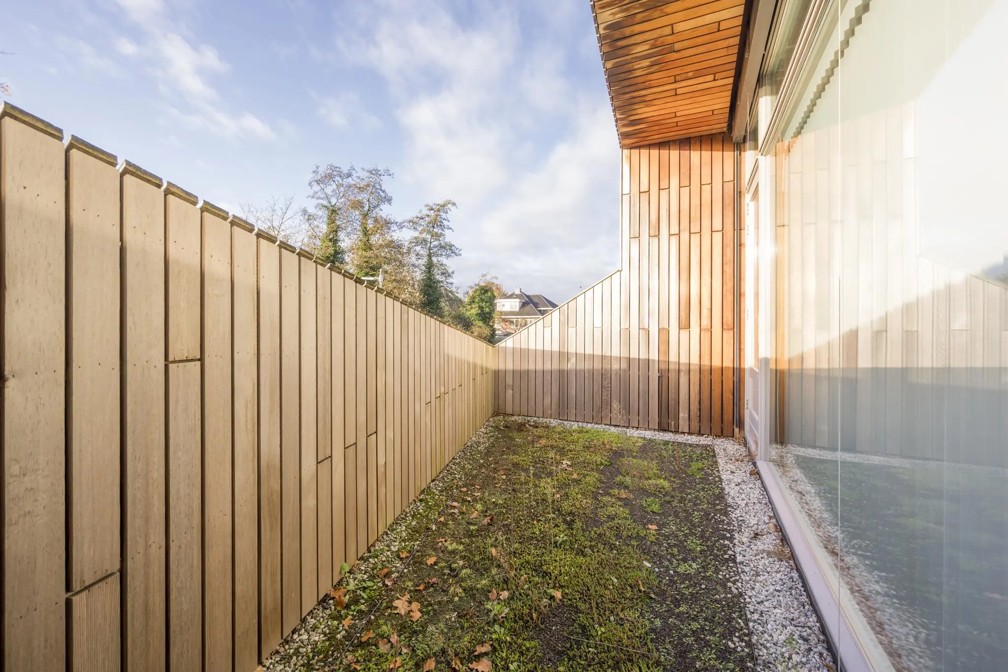 Kleine, smalle tuin met houten schutting en gevel aan de Vogelvlinderweg, met gras en grind langs het moderne gebouw.