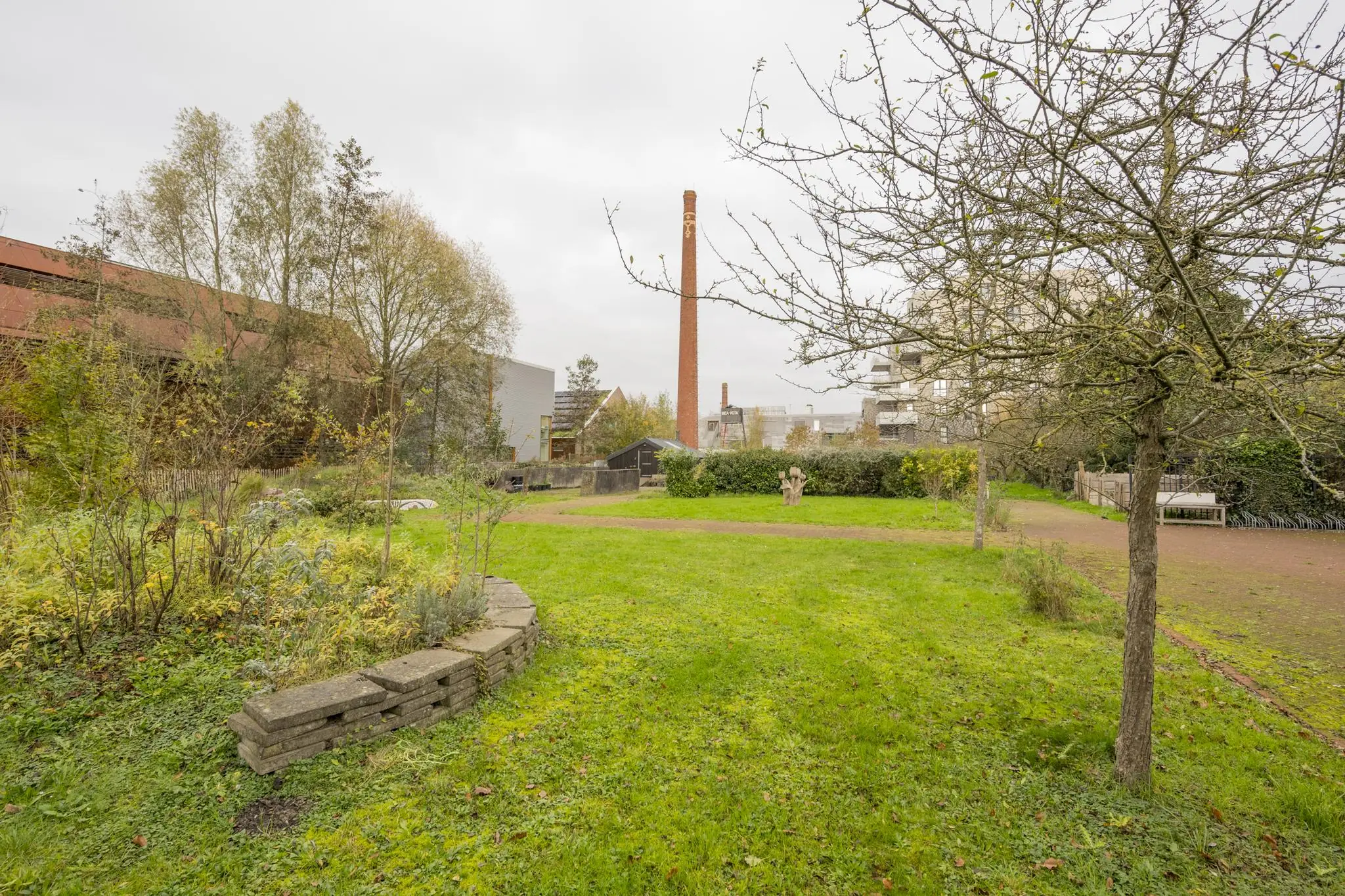 Groene binnentuin aan de Vogelvlinderweg met grasveld, bomen, planten en een hoge schoorsteen op de achtergrond.