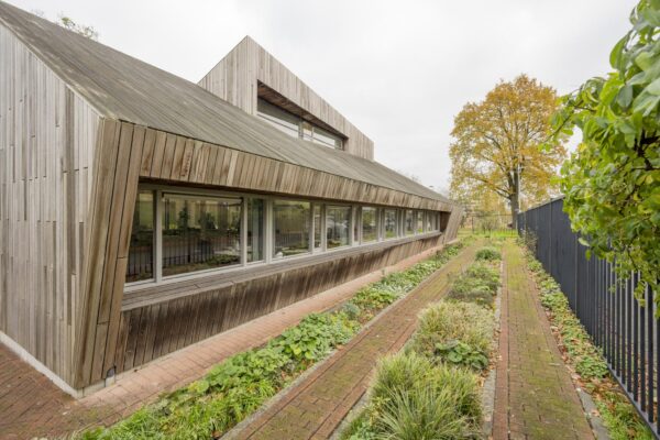Modern houten gebouw aan de Vogelvlinderweg met schuine gevel en lage ramen, naast een smal tuinpad met beplanting.