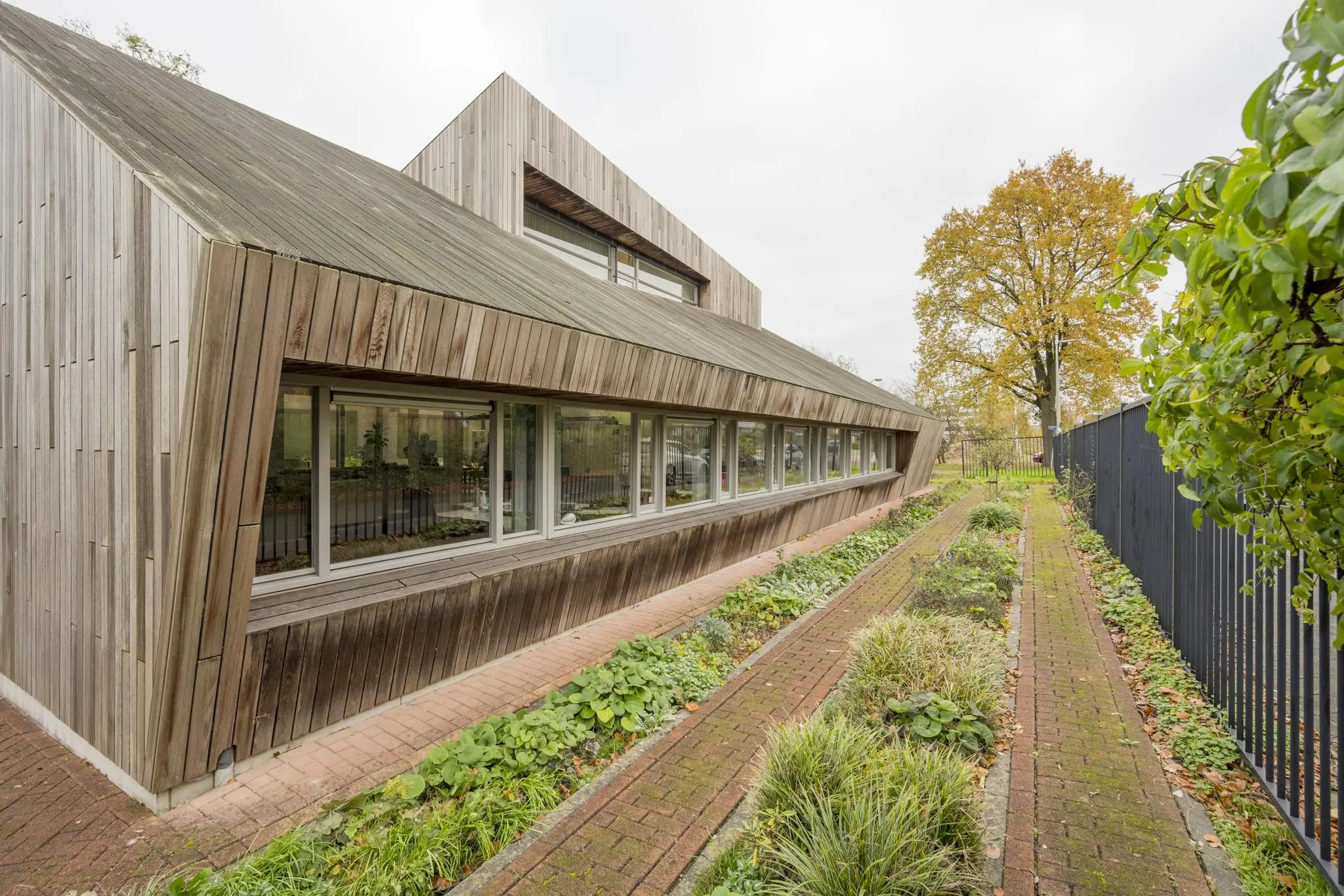 Modern houten gebouw aan de Vogelvlinderweg met schuine gevel en lage ramen, naast een smal tuinpad met beplanting.