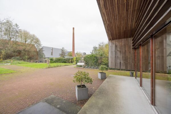 Binnenplaats met rode bestrating, plantenbakken en houten gevel aan de Vogelvlinderweg, met op de achtergrond een hoge bakstenen schoorsteen.