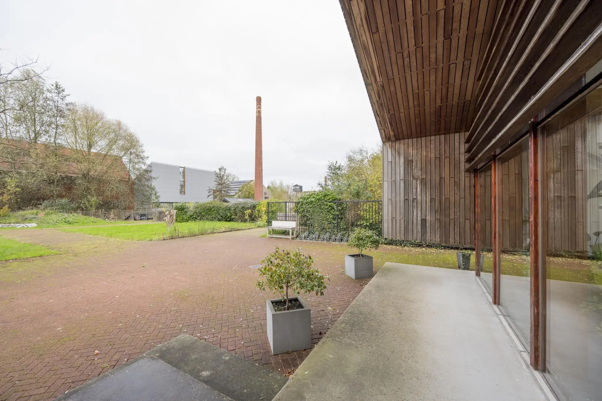 Binnenplaats met rode bestrating, plantenbakken en houten gevel aan de Vogelvlinderweg, met op de achtergrond een hoge bakstenen schoorsteen.