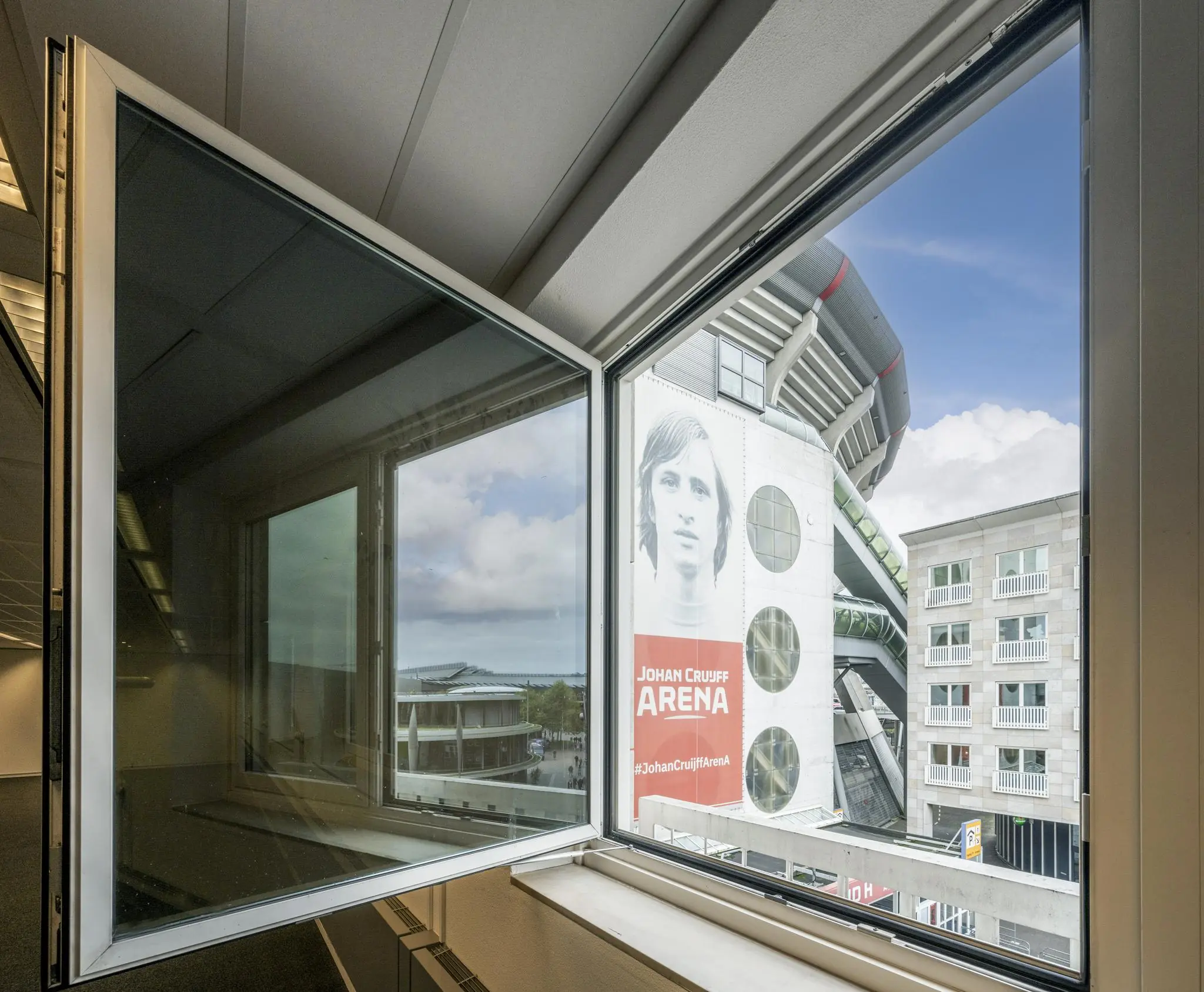 Uitzicht vanuit een open raam op de Johan Cruijff ArenA met een grote afbeelding van Johan Cruijff op de buitenmuur.