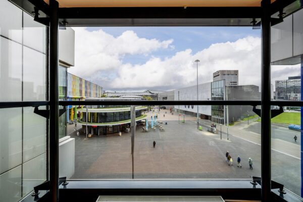 Uitzicht vanaf een raam op de Johan Cruijff Boulevard met moderne gebouwen, wandelend publiek en een plein.