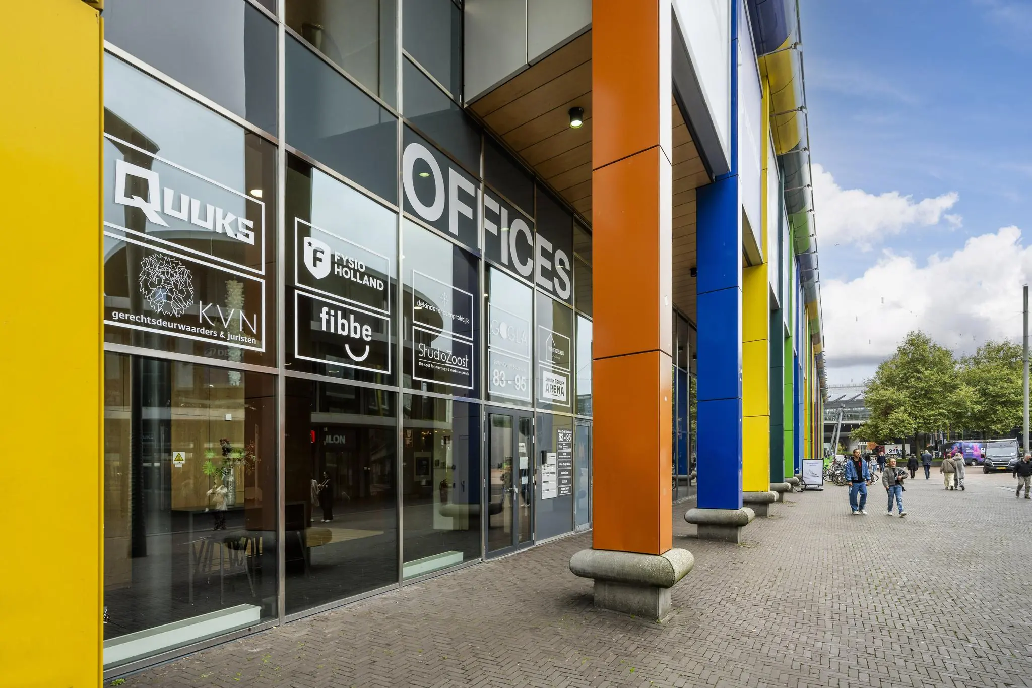 Ingang van een kantoorpand aan de Johan Cruijff Boulevard met kleurrijke zuilen en passerende voetgangers.