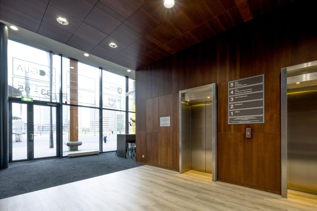 Moderne entreehal aan de Johan Cruijff Boulevard met liften, houten wandafwerking en zicht op kantoorgebouwen buiten.