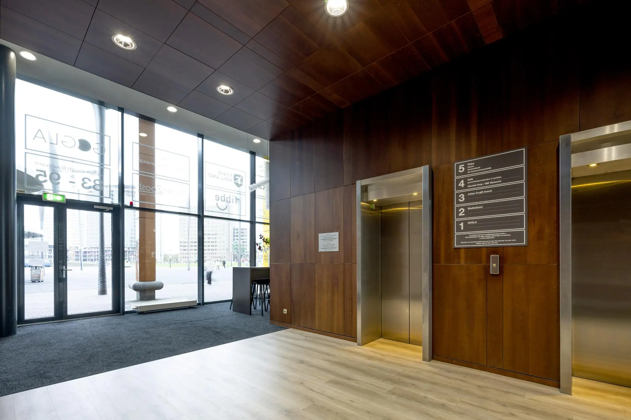 Moderne entreehal aan de Johan Cruijff Boulevard met liften, houten wandafwerking en zicht op kantoorgebouwen buiten.