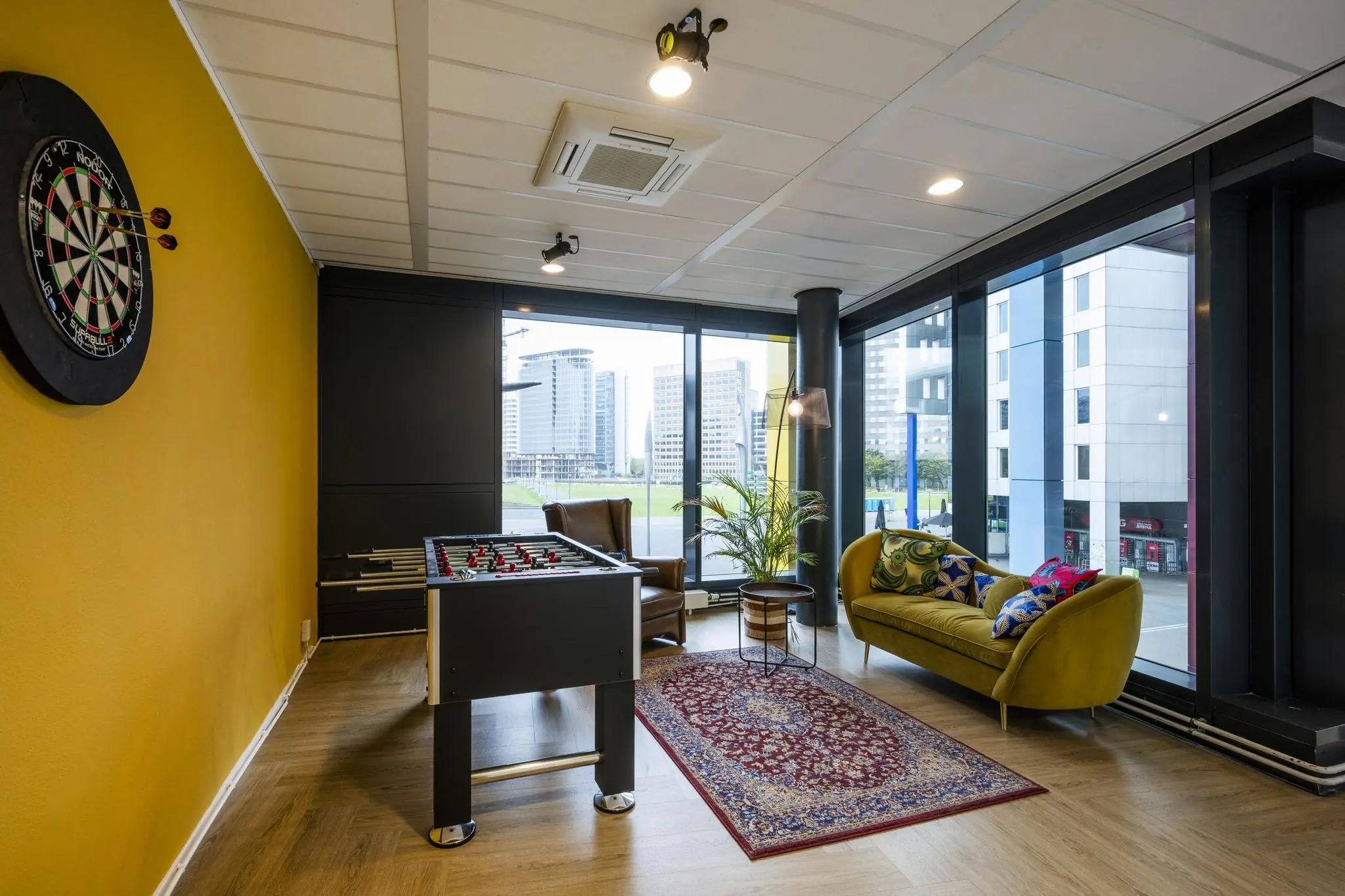 Speelse loungehoek aan de Johan Cruijff Boulevard met tafelvoetbaltafel, dartbord en uitzicht op moderne kantoorgebouwen.