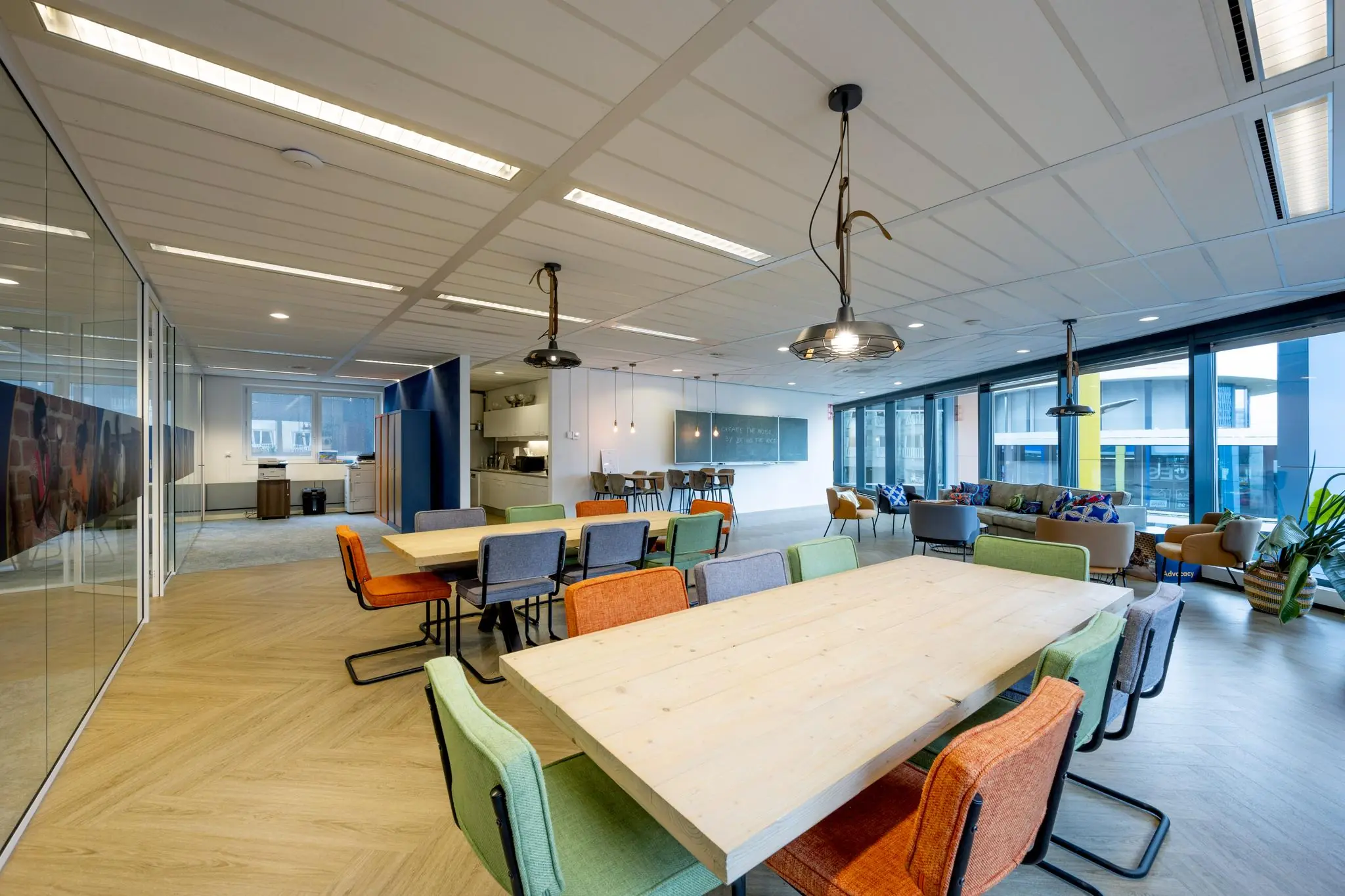 Moderne kantoorruimte aan de Johan Cruijff Boulevard met kleurrijke stoelen, houten tafels en een zithoek bij grote ramen.