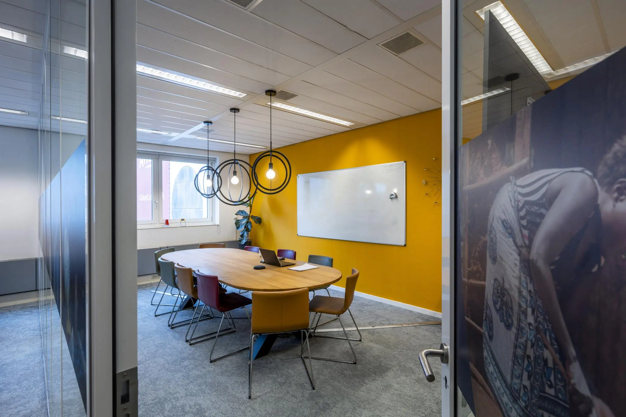 Vergaderruimte aan de Johan Cruijff Boulevard met ovale tafel, kleurrijke stoelen, gele accentmuur en modern hanglampontwerp.