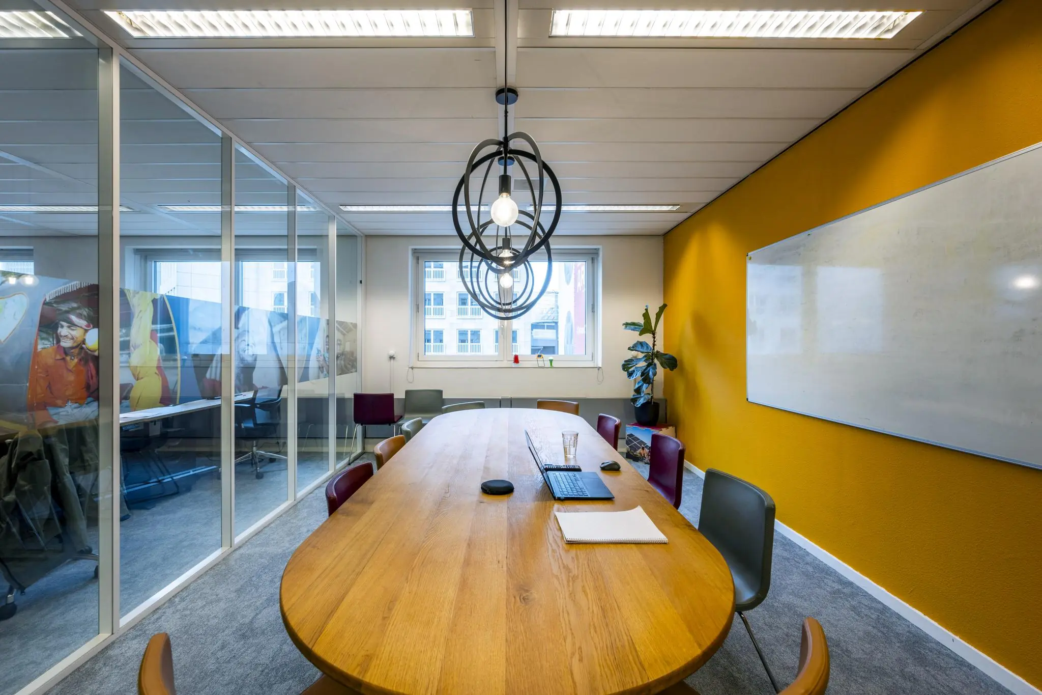 Vergaderkamer aan de Johan Cruijff Boulevard met een ovale houten tafel, kleurrijke stoelen, glazen wand, laptop en whiteboard.