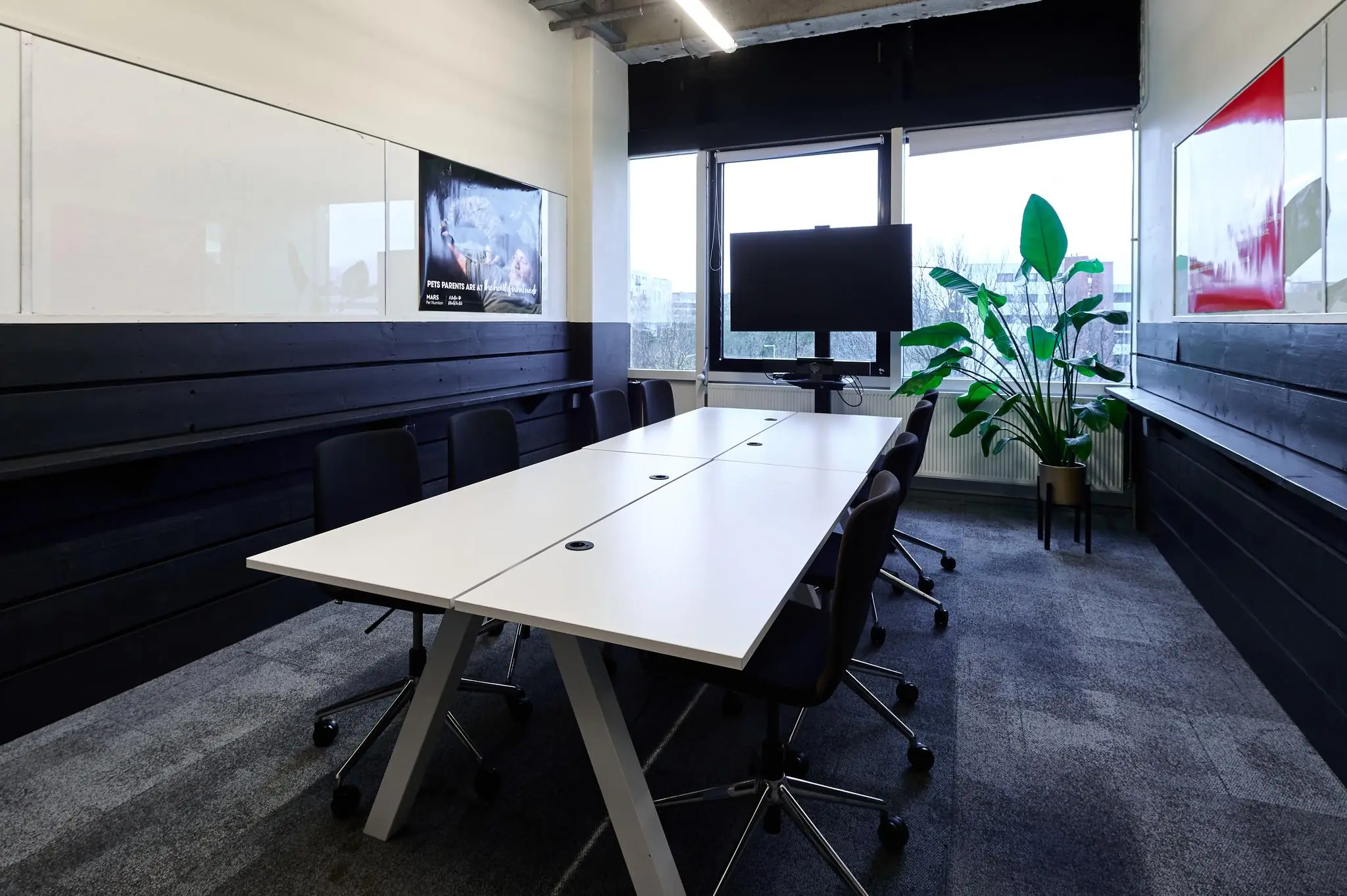 Vergaderruimte aan de Johan Huizingalaan met een lange witte tafel, zwarte stoelen, een scherm en grote plant bij het raam.