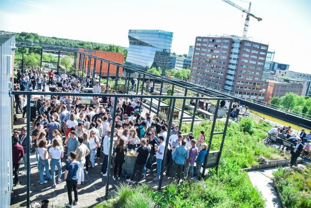 Drukke borrel op een zonnig dakterras aan de Johan Huizingalaan met uitzicht op moderne kantoorgebouwen.