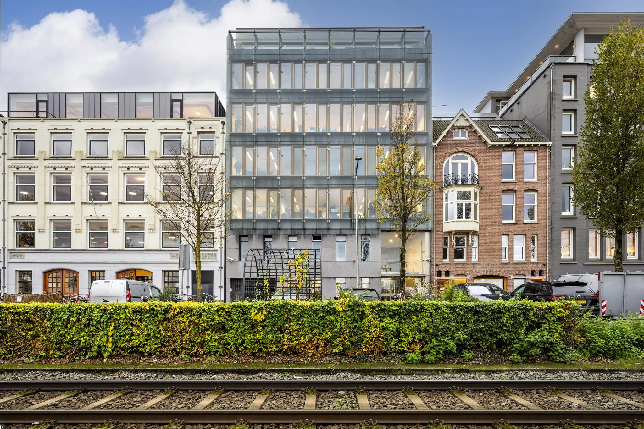 Kantoorpand met een moderne glazen gevel aan de De Ruijterkade in Amsterdam, geflankeerd door klassieke gebouwen, met daarvoor spoorrails en een groene haag.