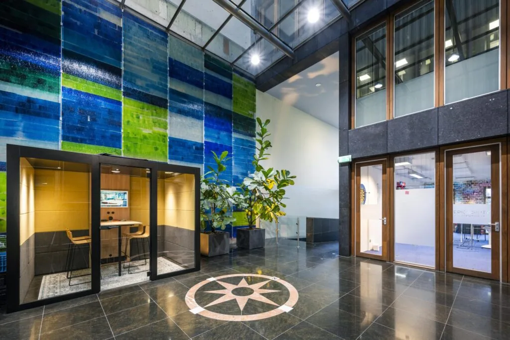 Moderne lobby van een kantoorgebouw aan de De Ruijterkade met glaswanden, kleurrijke wanddecoratie, planten en een geluidsdichte vergaderruimte.