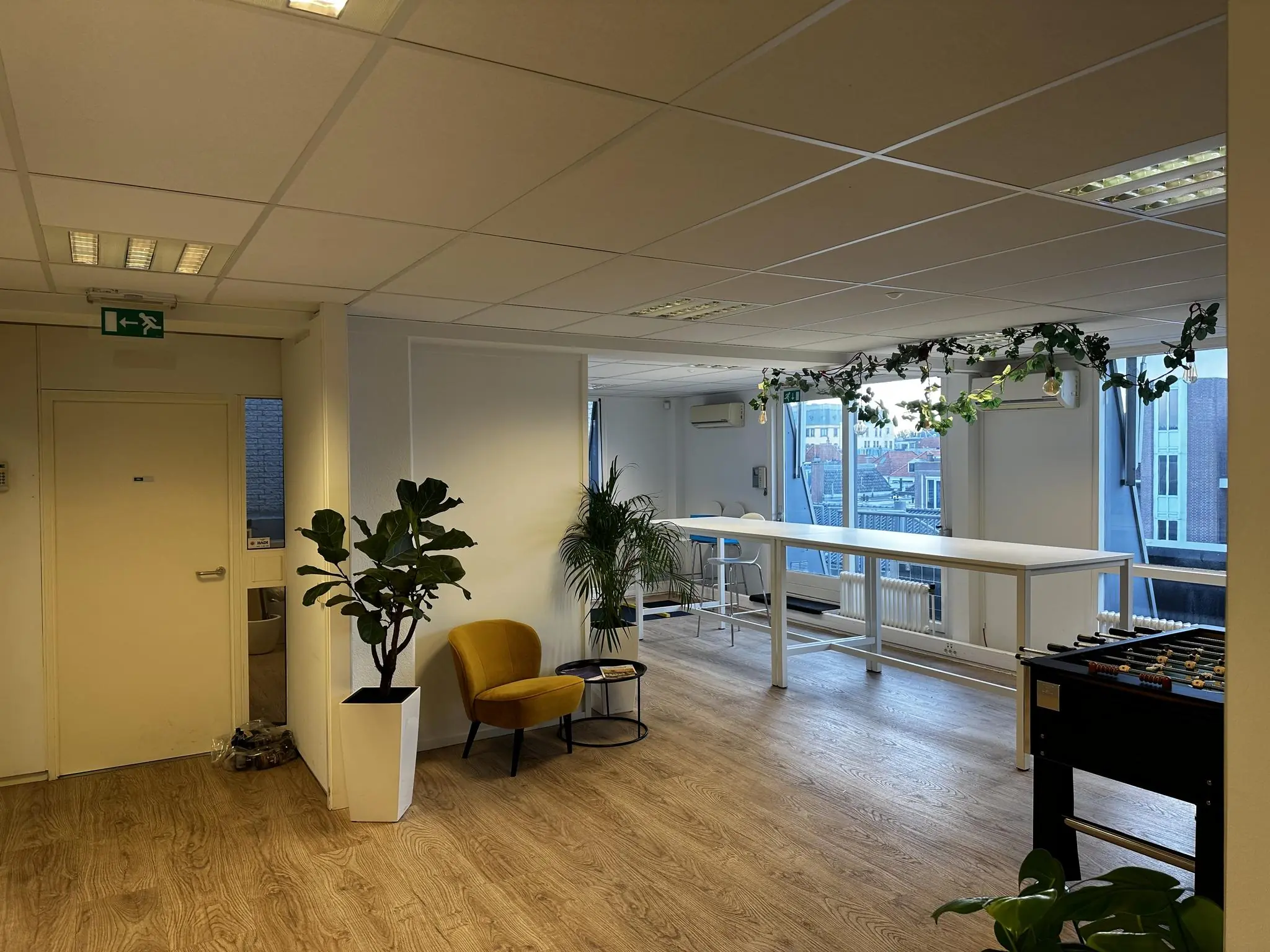 Modern kantoorinterieur aan de Koninginnegracht met hoge tafels, planten, een voetbaltafel en grote ramen met uitzicht op de stad.