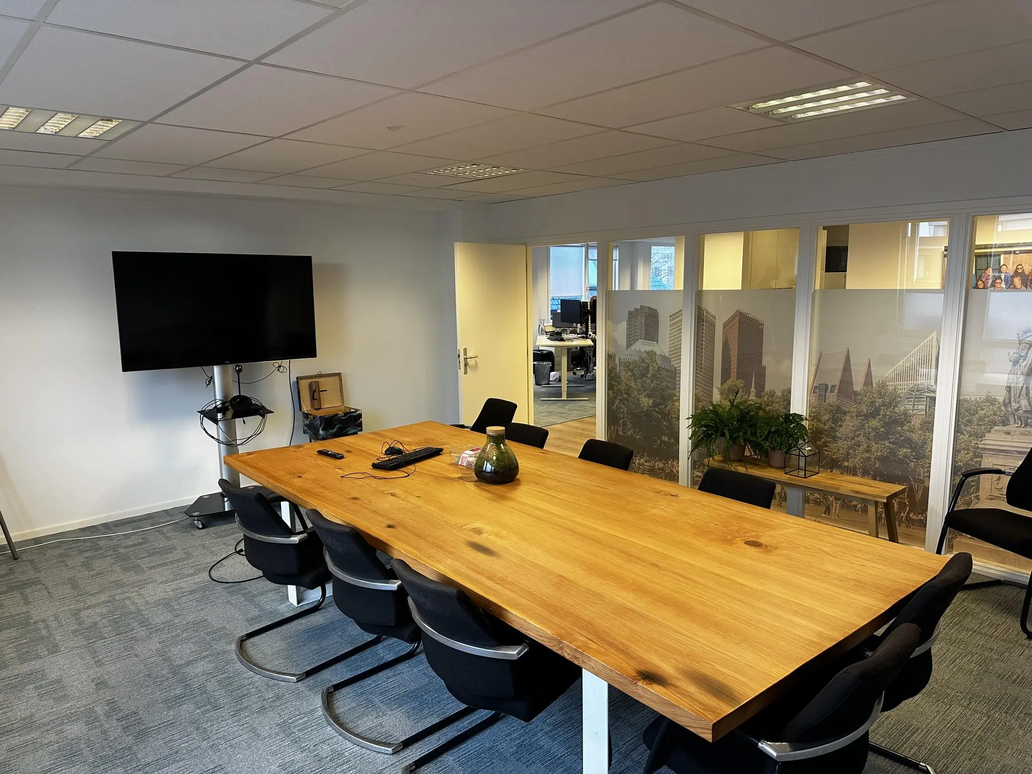 Vergaderruimte aan de Koninginnegracht met houten tafel, zwarte stoelen, televisie en glazen wand met stadsafbeeldingen.