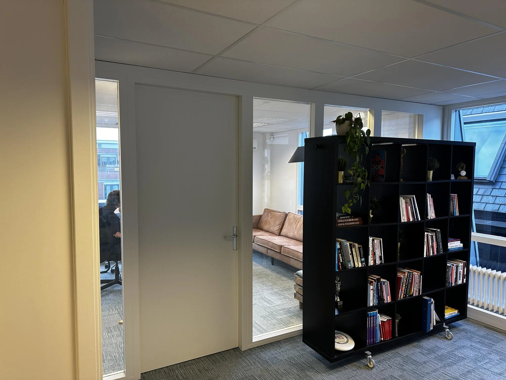 Moderne kantoorruimte aan de Koninginnegracht met glazen wanden, een bruine bank en een zwarte boekenkast vol boeken en planten.