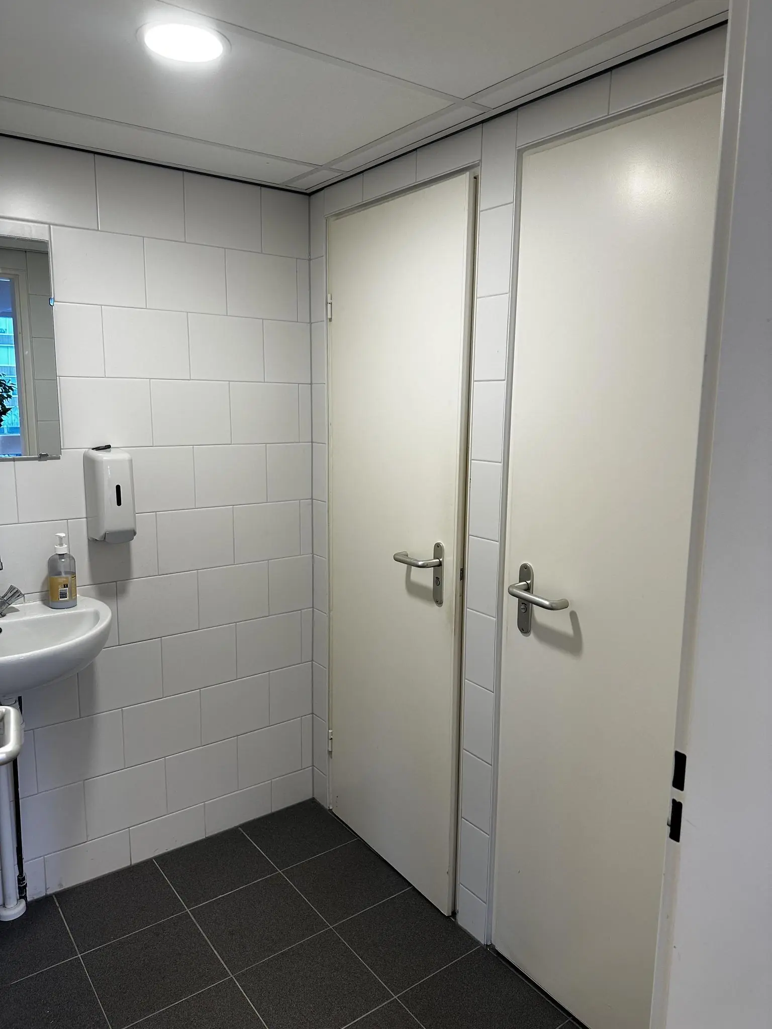 Wit betegelde toiletruimte aan de Koninginnegracht met twee gesloten deuren en een wasbak met zeepdispenser.