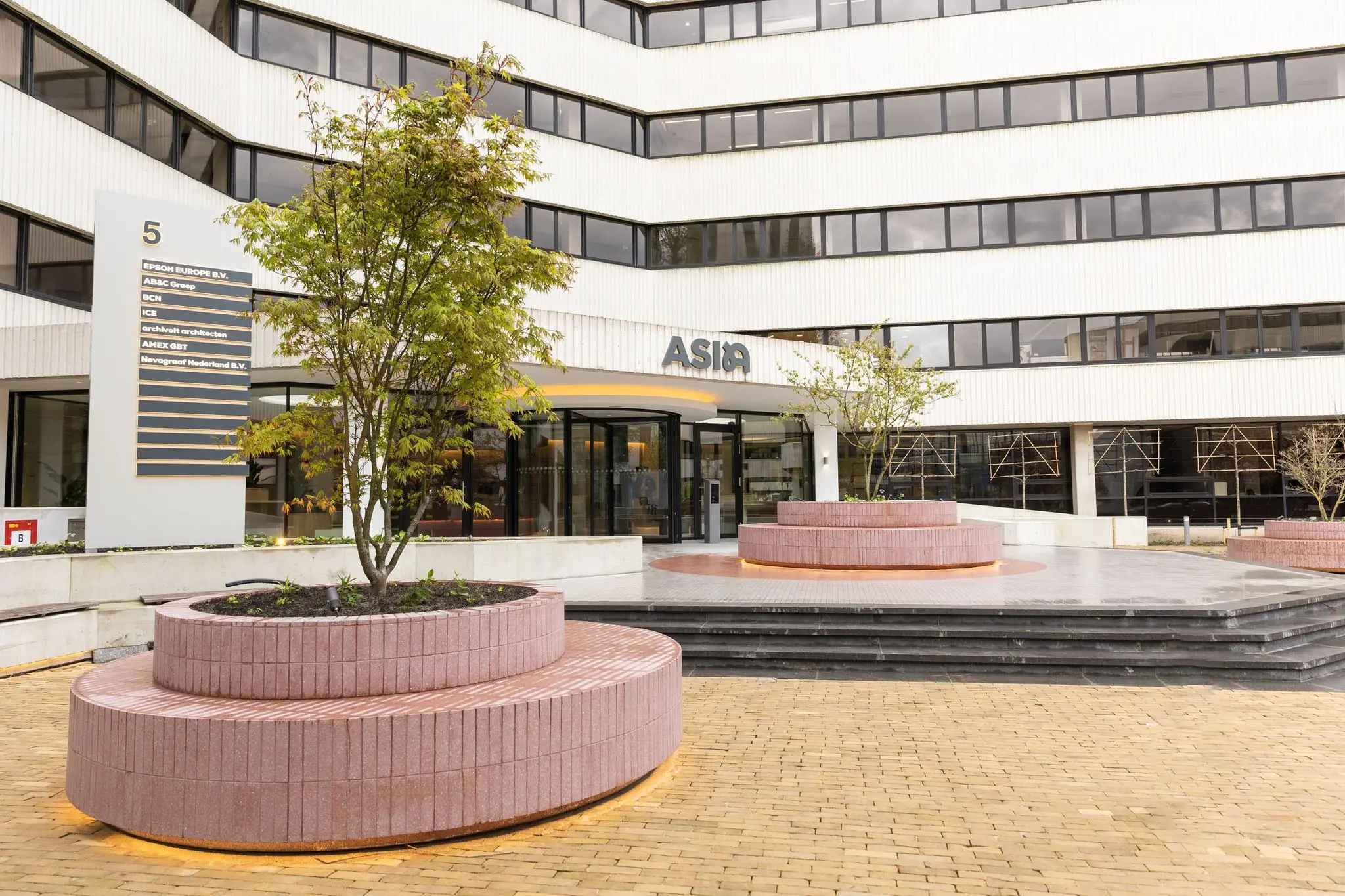 De hoofdingang van het kantoorgebouw aan de Hoogoorddreef 5 in Amsterdam met bedrijfsnamenlijst en ronde plantenbakken op het plein.