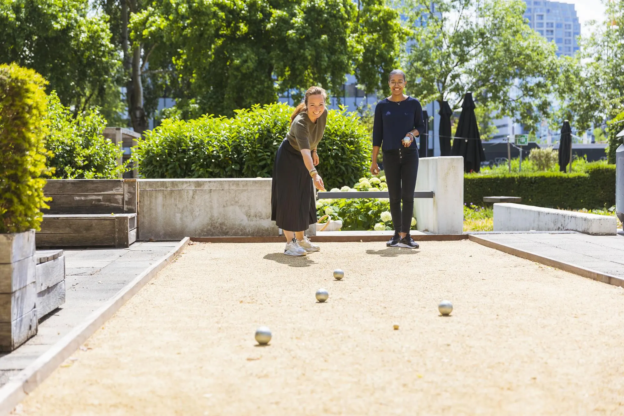 Twee vrouwen spelen jeu de boules op een zonnige dag aan de Hoogoorddreef in een groene, stedelijke omgeving.