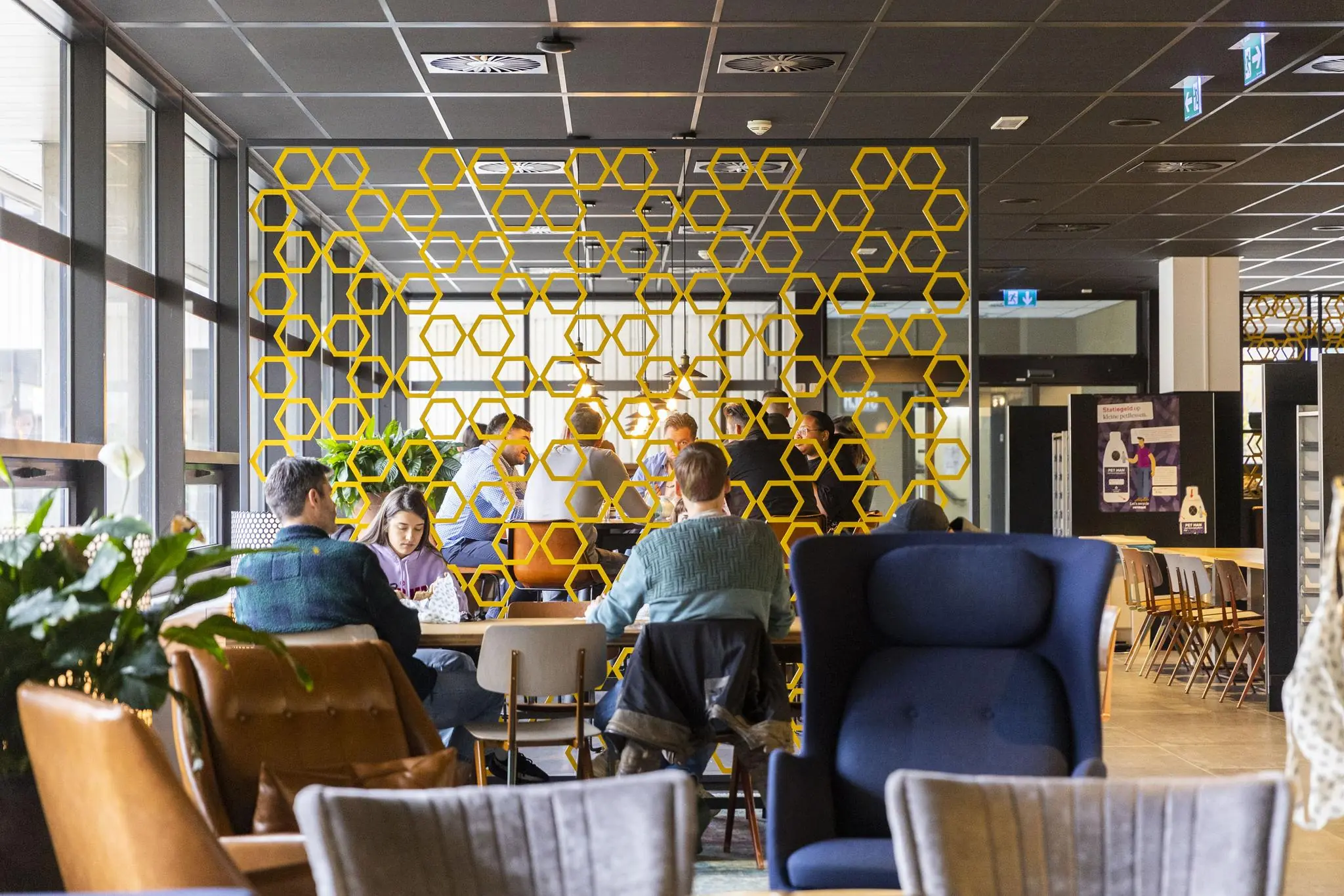 Mensen werken en overleggen in een modern ingerichte ruimte aan de Hoogoorddreef, gescheiden door een decoratief geel honingraat-scherm.