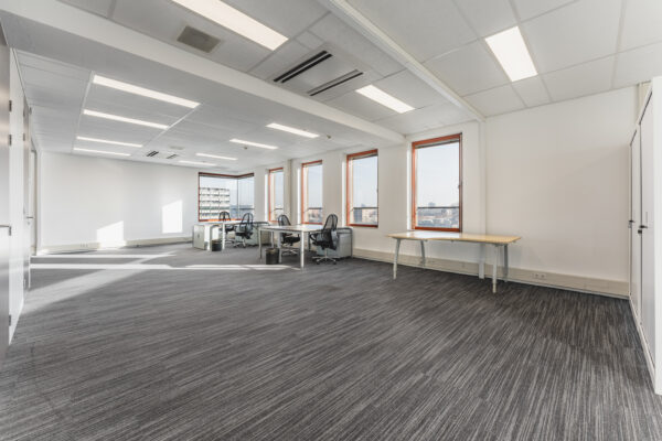 Een ruime, lege kantoorruimte met bureaus, stoelen en grote ramen.