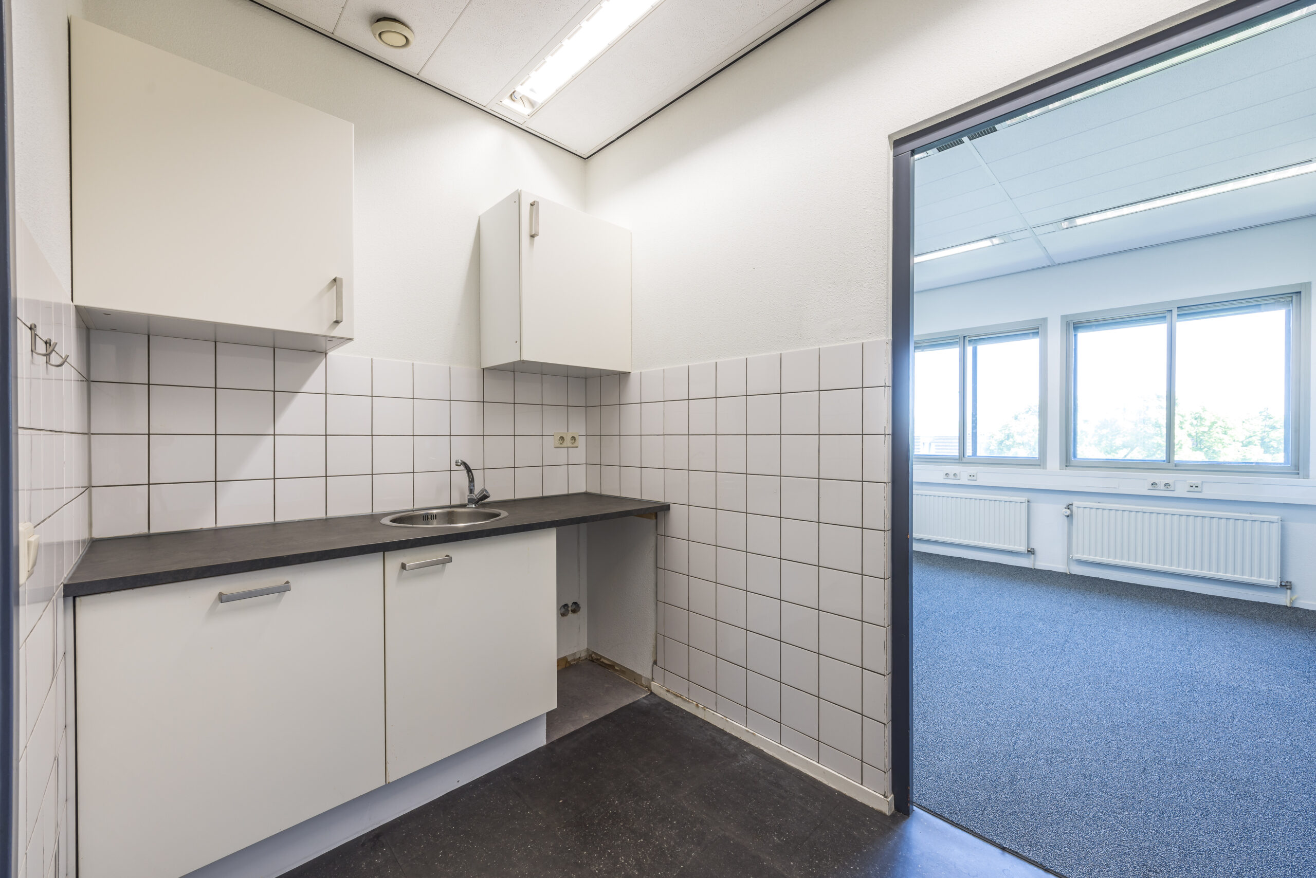 Kleine kantoorruimte met keukenblok en betegelde muur, naast een kamer met tapijt en grote ramen.