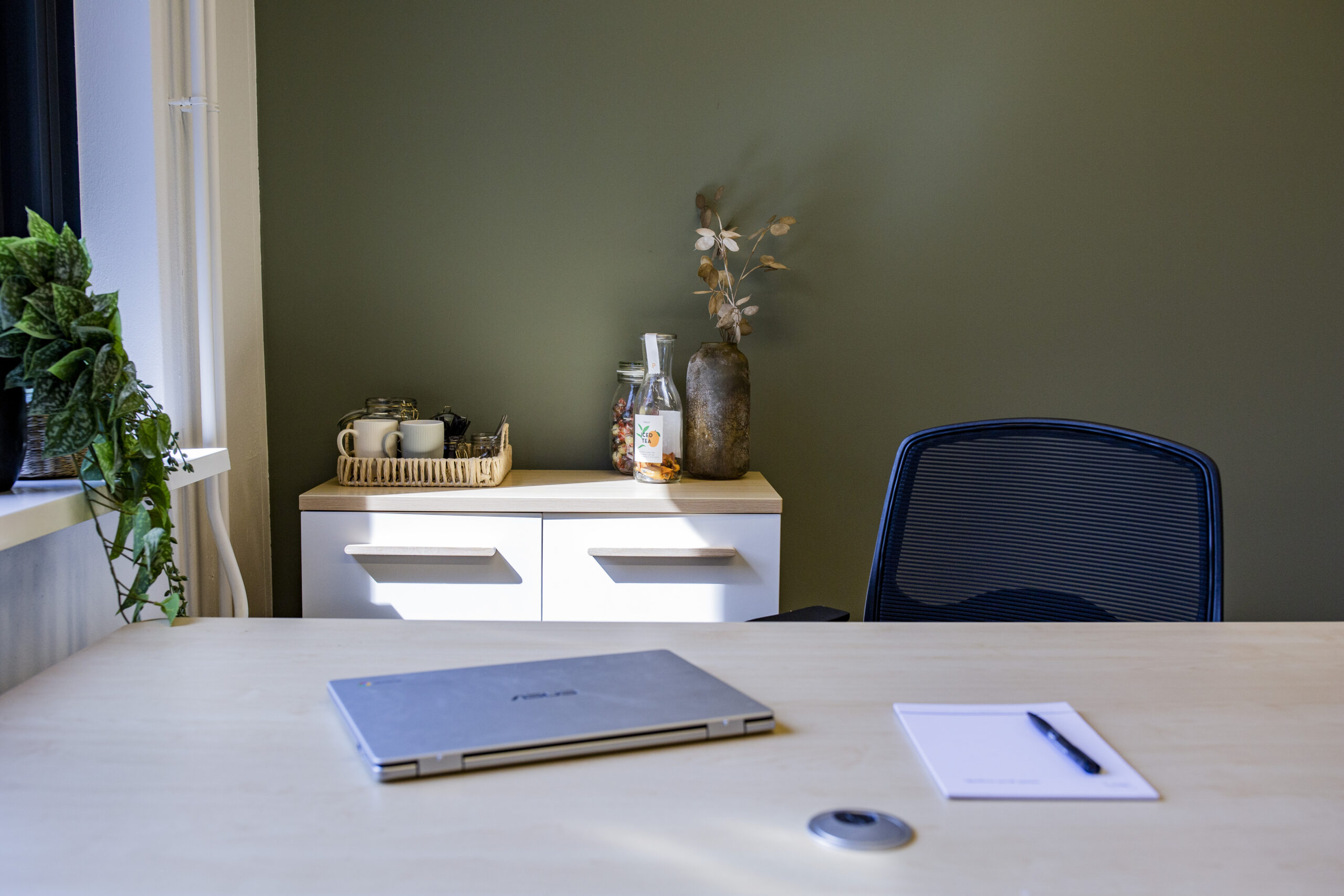 Een modern kantoor met een bureau, laptop, stoel en kast met decoraties en een dienblad met koffiekopjes tegen een groene muur.