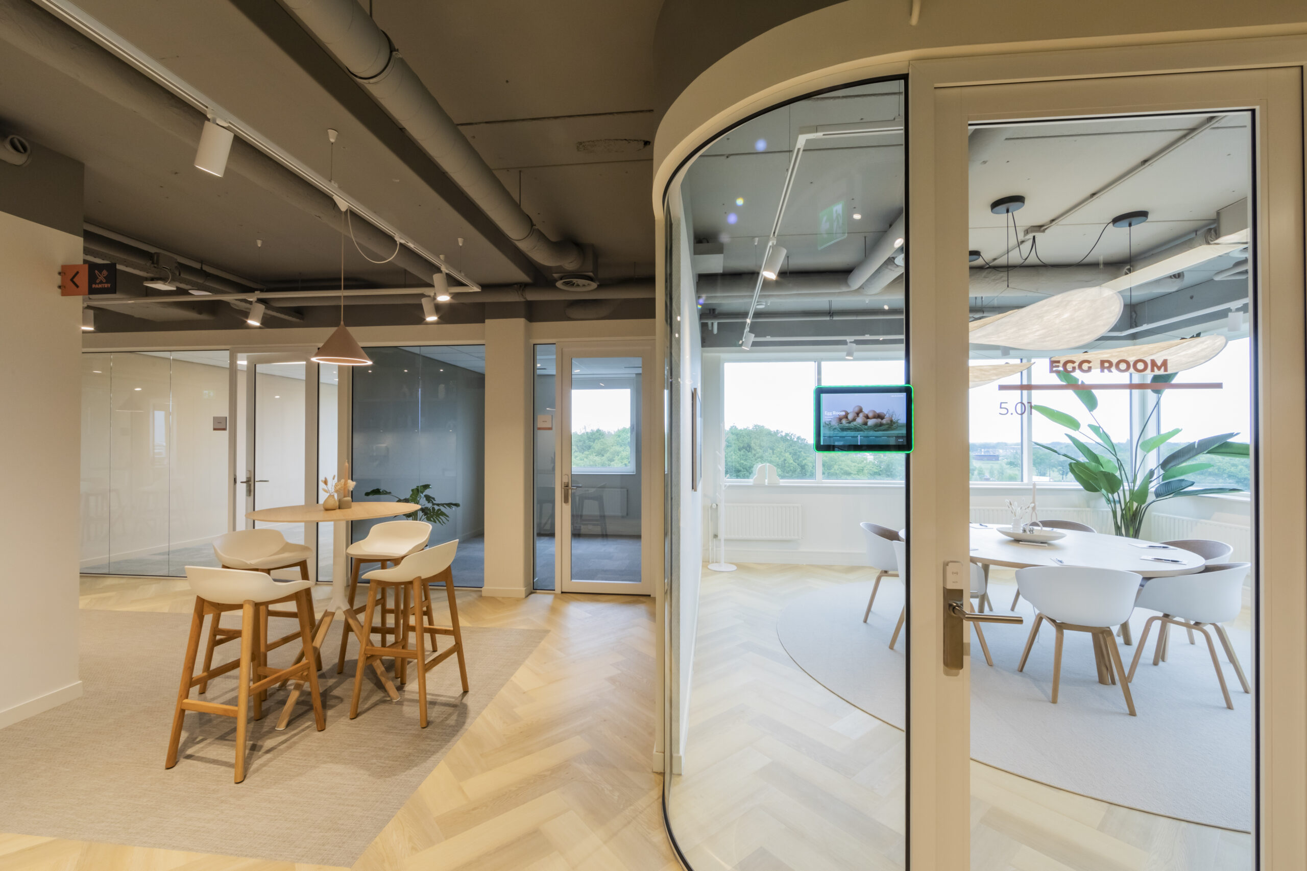 Een modern kantoorinterieur met houten vloer, ronde glazen vergaderruimte genaamd "Egg Room," en hoge stoelen rond een tafel.