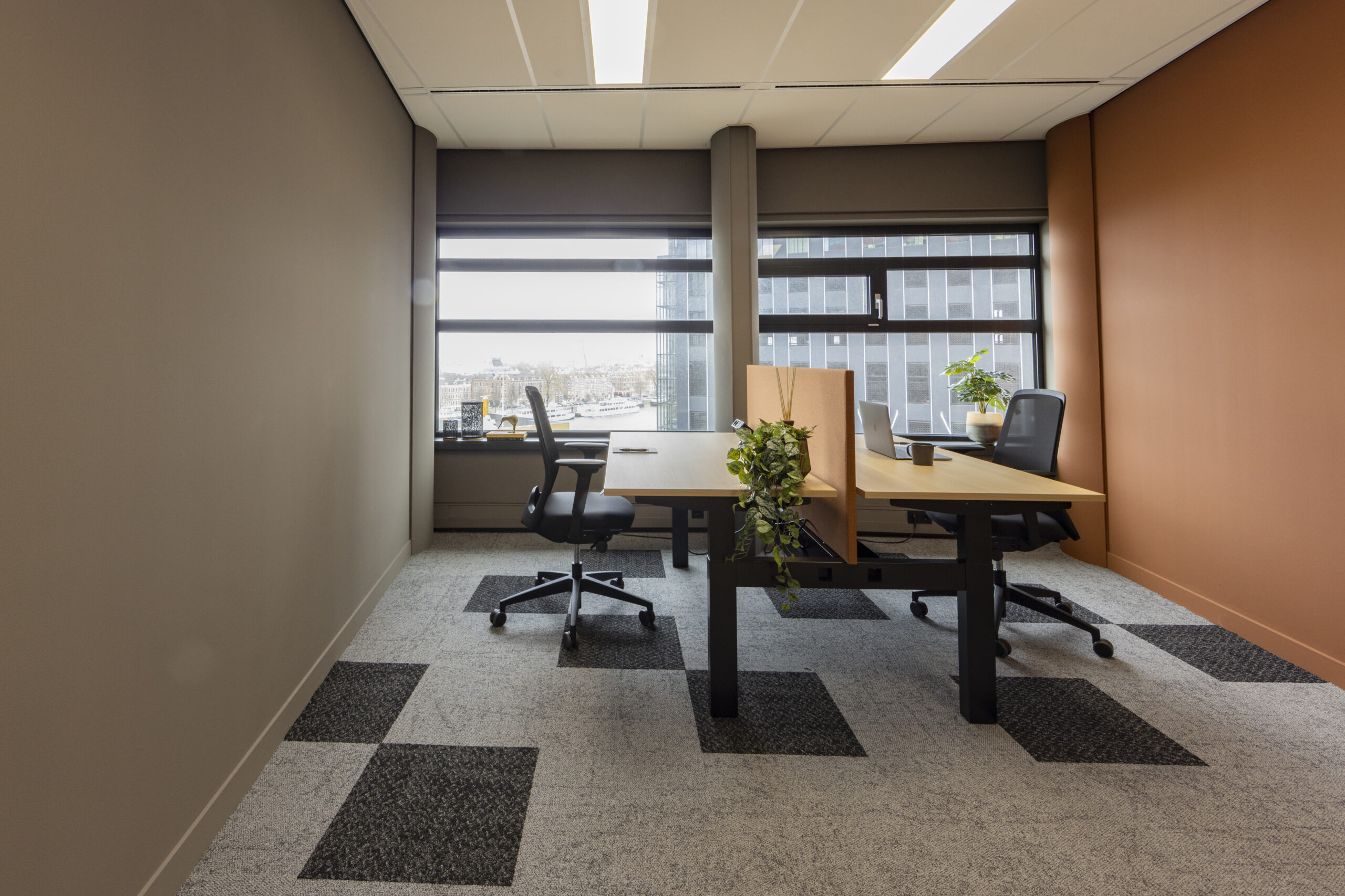 Een modern kantoor in Rotterdam met een groot raam, twee bureaus, planten en bureaustoelen.