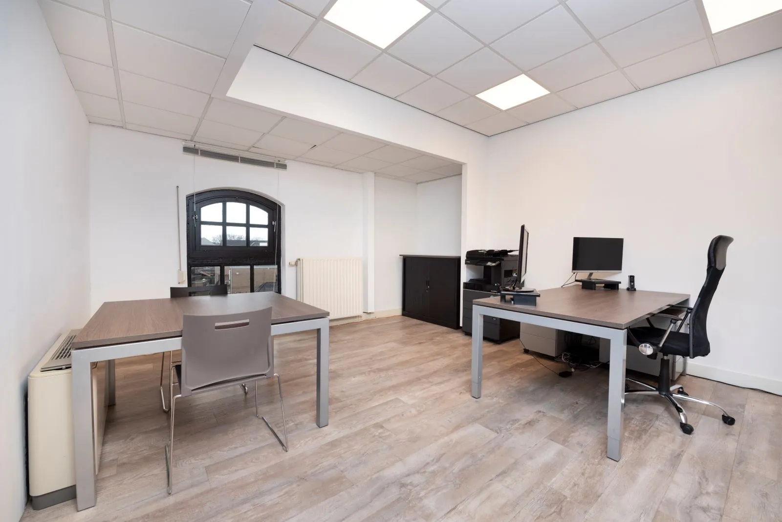 Modern kantoor met twee bureaus, een computeropstelling en een zwart boograam in ruimte 210 Suiker.