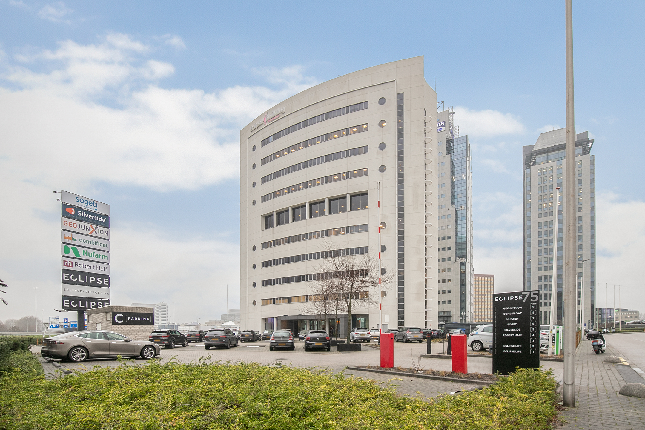 Een modern kantoorgebouw genaamd Rivium Quadrant met een parkeergarage en bedrijfsnamen op borden.