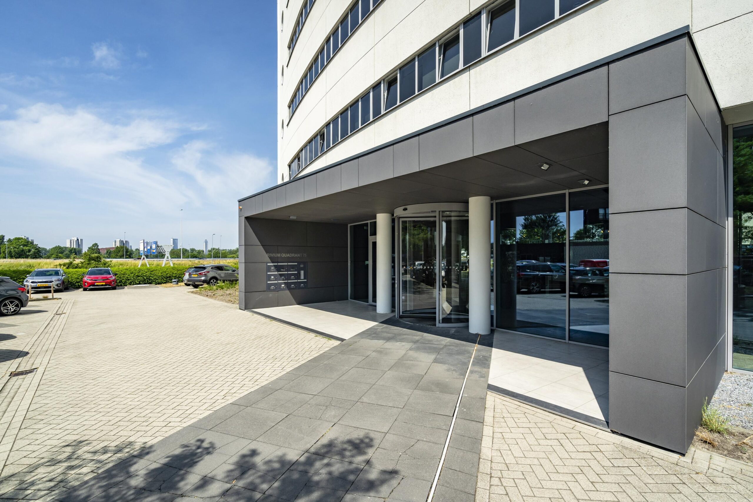 Vooraanzicht van het Rivium Quadrant-kantoorgebouw met een roterende entree en parkeerplaats met auto's.