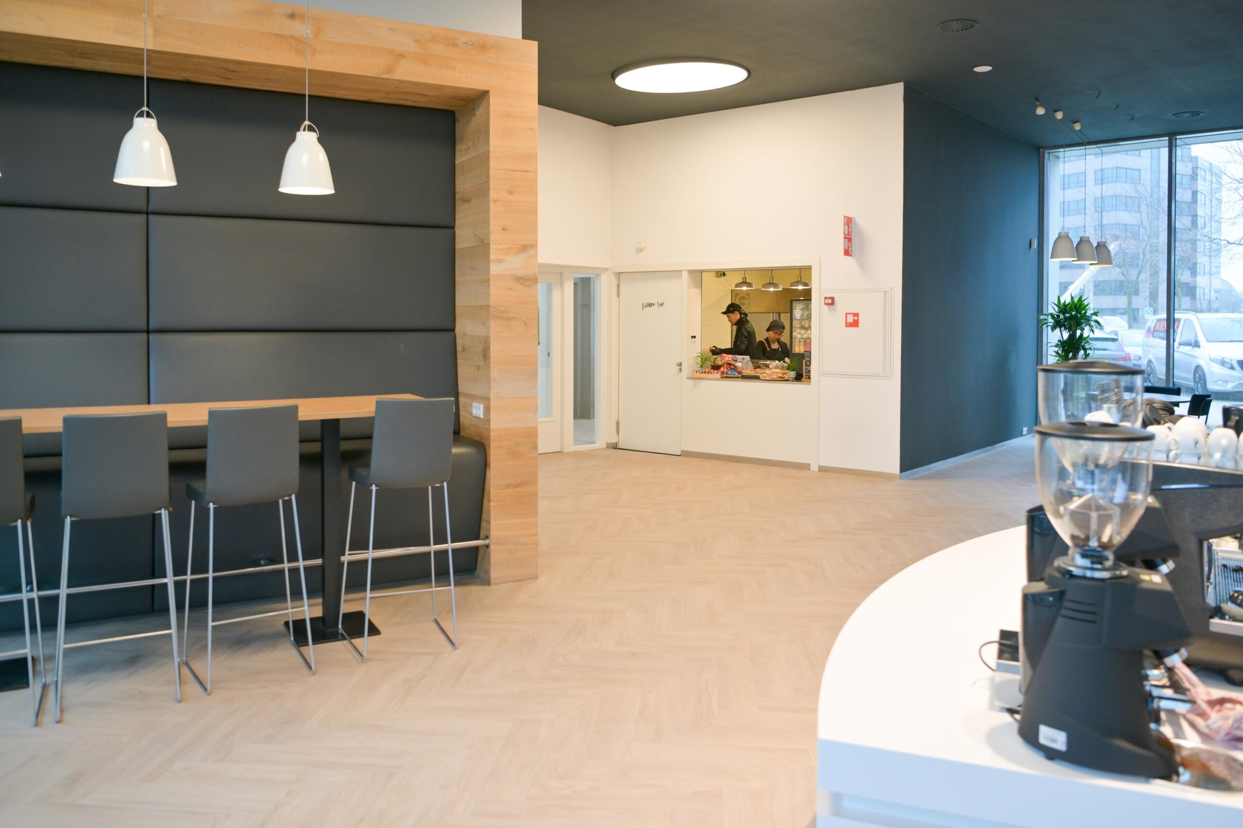 Moderne kantineruimte met hoge stoelen aan een bar, een koffiezetapparaat en een kleine keuken in de achtergrond.